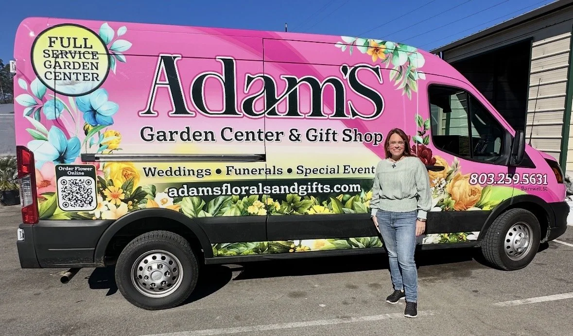 A woman stands in front of a pink floral Adams Garden Center & Gift Shop delivery van parked in a lot.