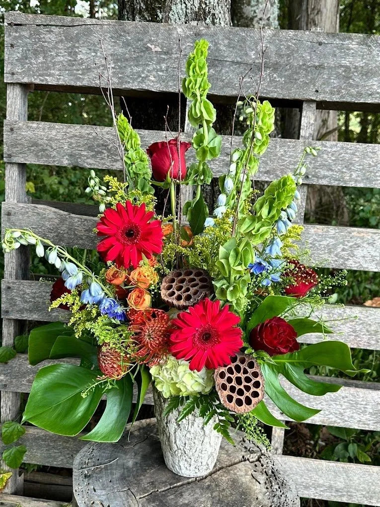A colorful flower arrangement in a white vase featuring red gerbera daisies, red roses, blue delphiniums, green foliage, and lotus seed pods, placed on a rustic wooden surface with a weathered wooden slat background.