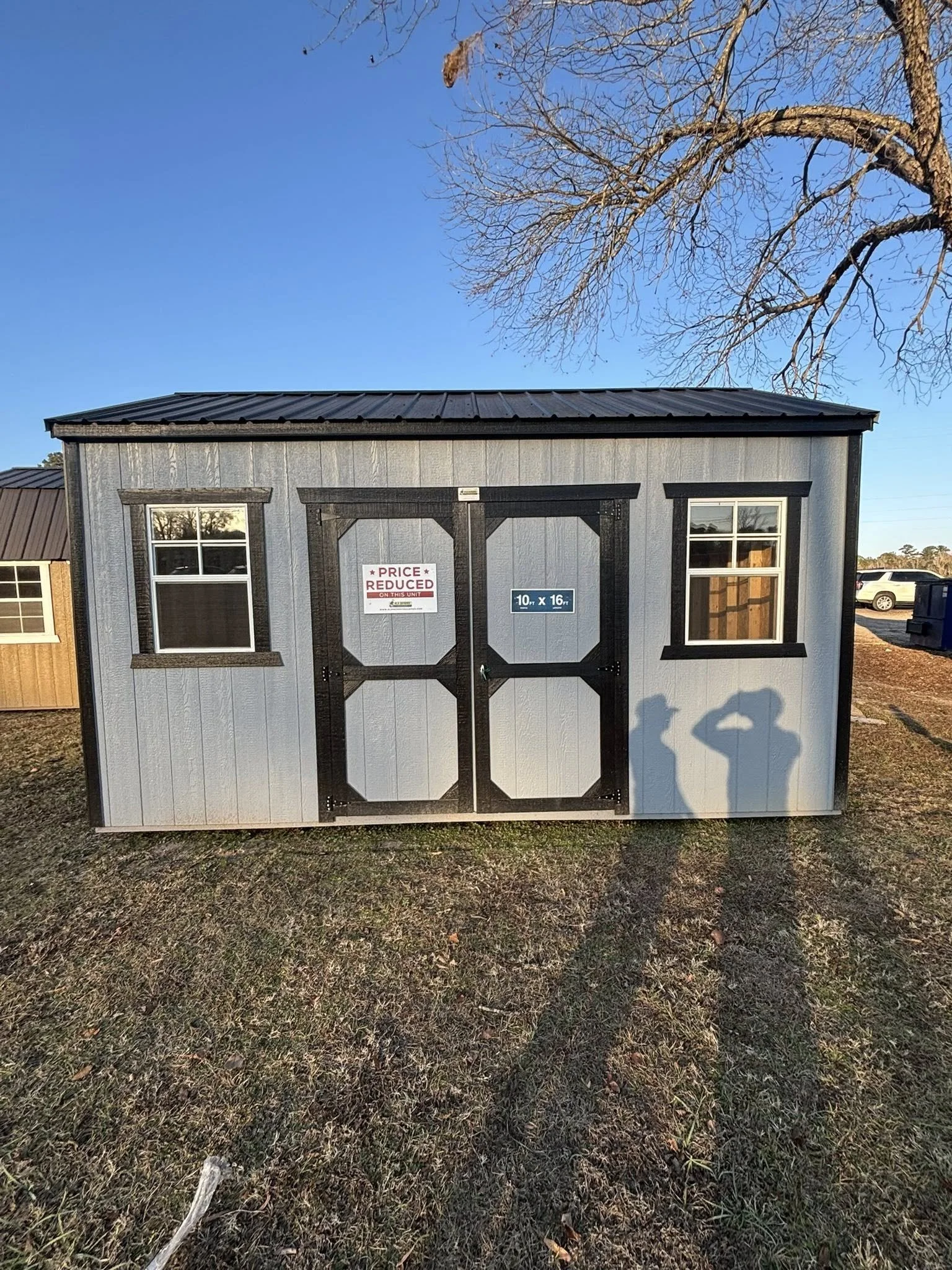 Small gray storage shed with black trim, two windows, and black doors, set outdoors on grassy ground. Shadows of two people taking the photo are visible on the shed. Sign on the shed indicating dimensions and a red sign stating 'Price Reduced.'