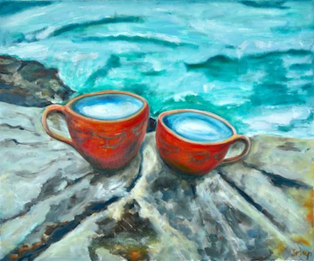Two red ceramic coffee cups with blue liquid inside, placed on a rock near the water at a beach or lakeside. The background features turquoise water and rocky shoreline.