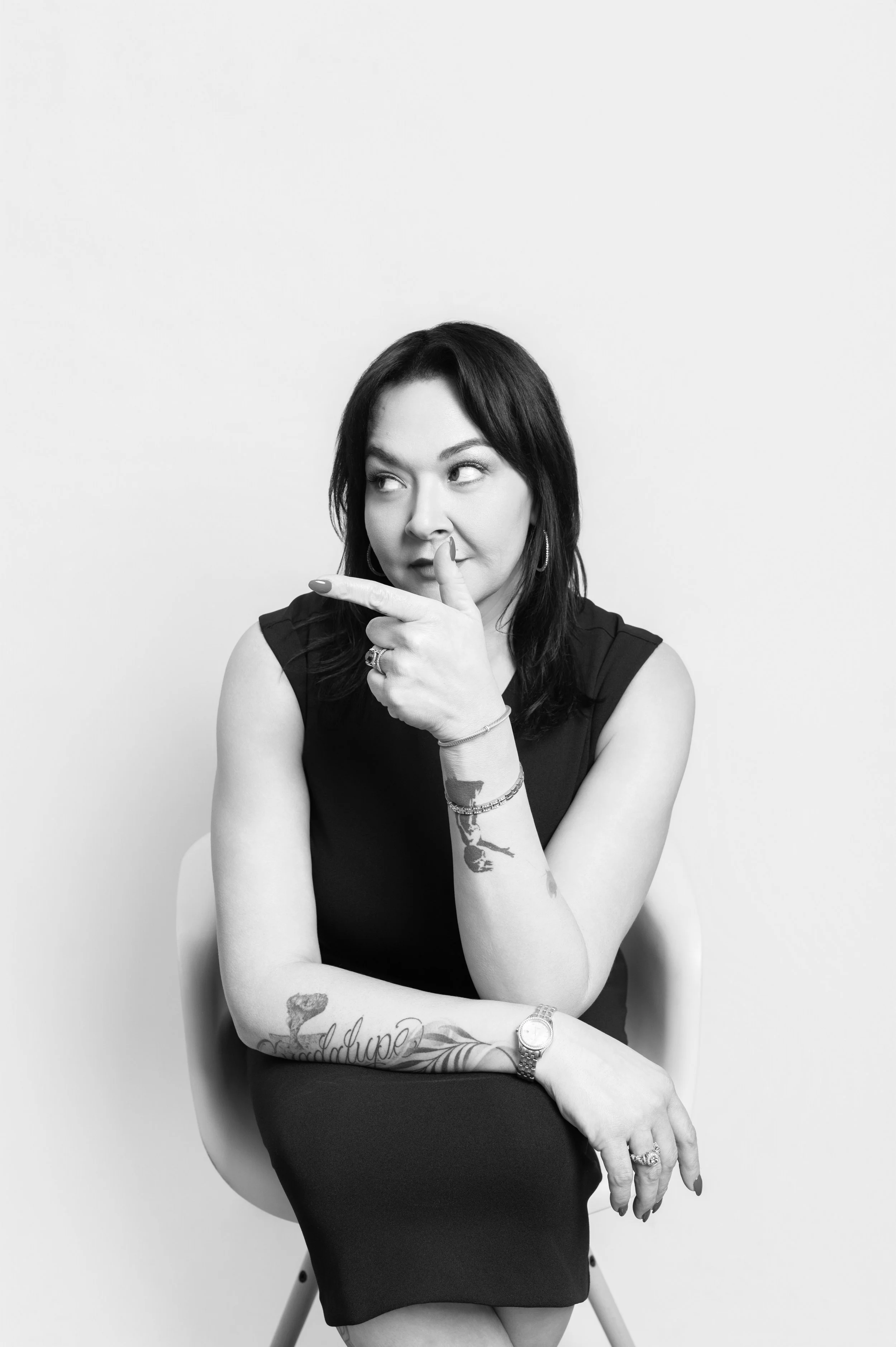 Black and white portrait of a woman with dark hair, tattoos on her left arm, wearing a sleeveless top, sitting on a chair, holding her chin with her index finger, and looking to her right.