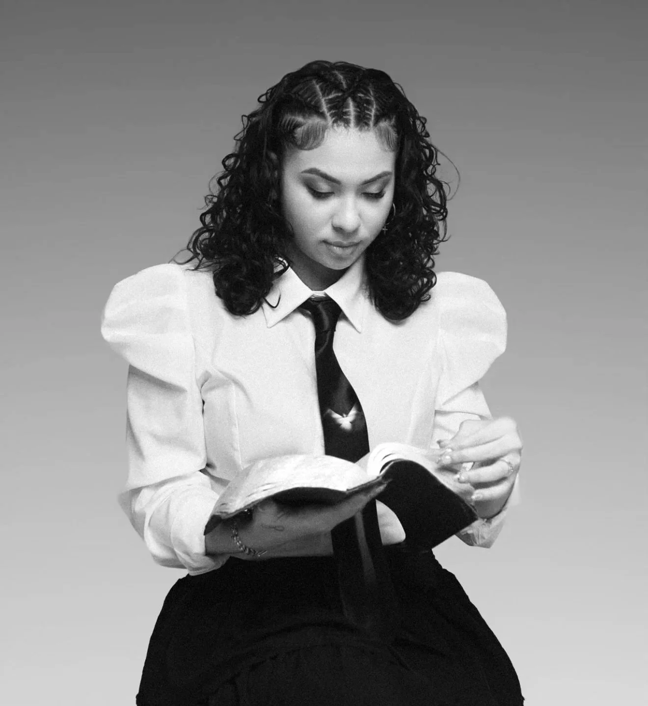 A young woman with curly hair wearing a white shirt, black tie, and dark skirt, sitting and reading a book.