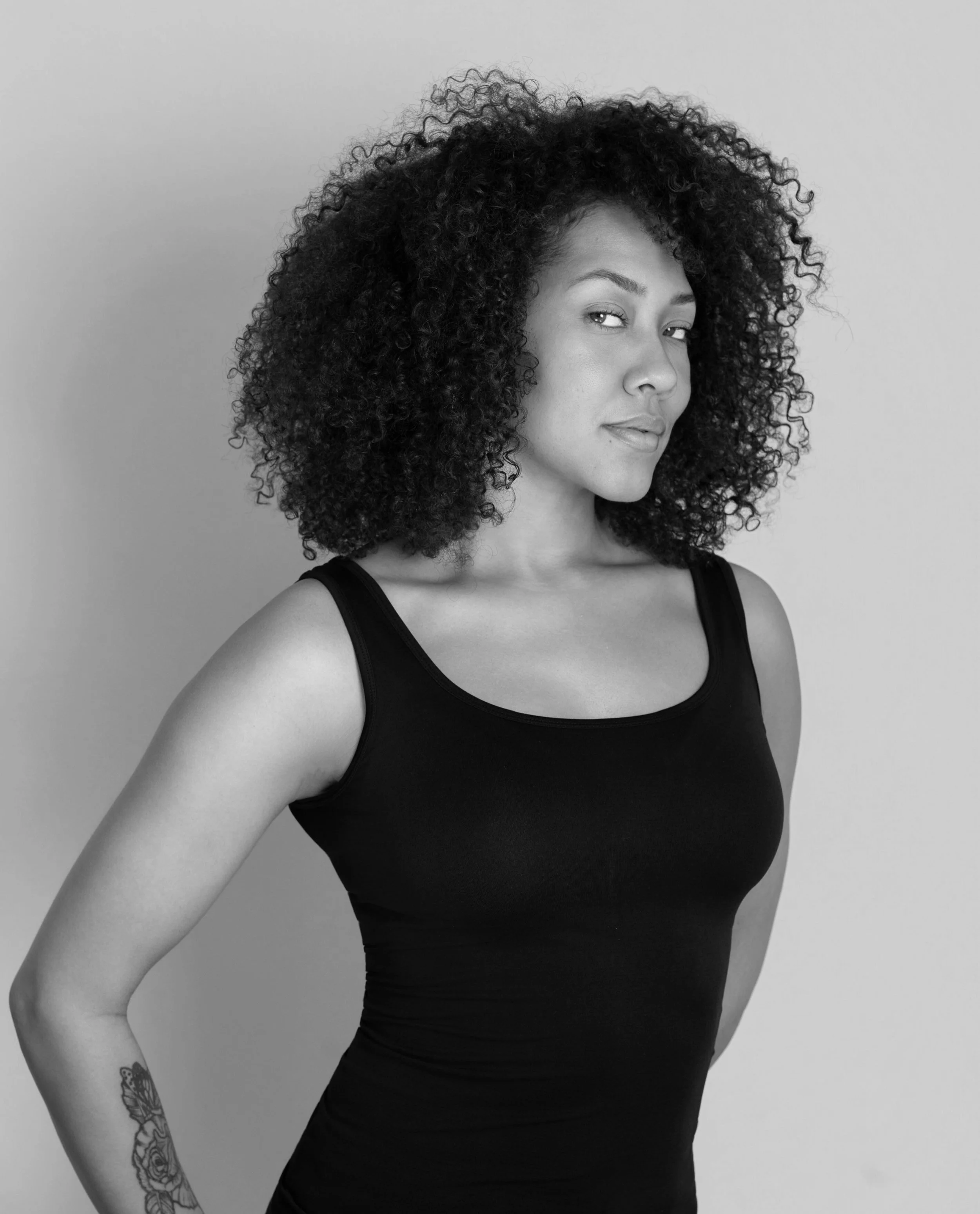 Black and white portrait of a woman with curly hair wearing a sleeveless black top, standing against a light background.