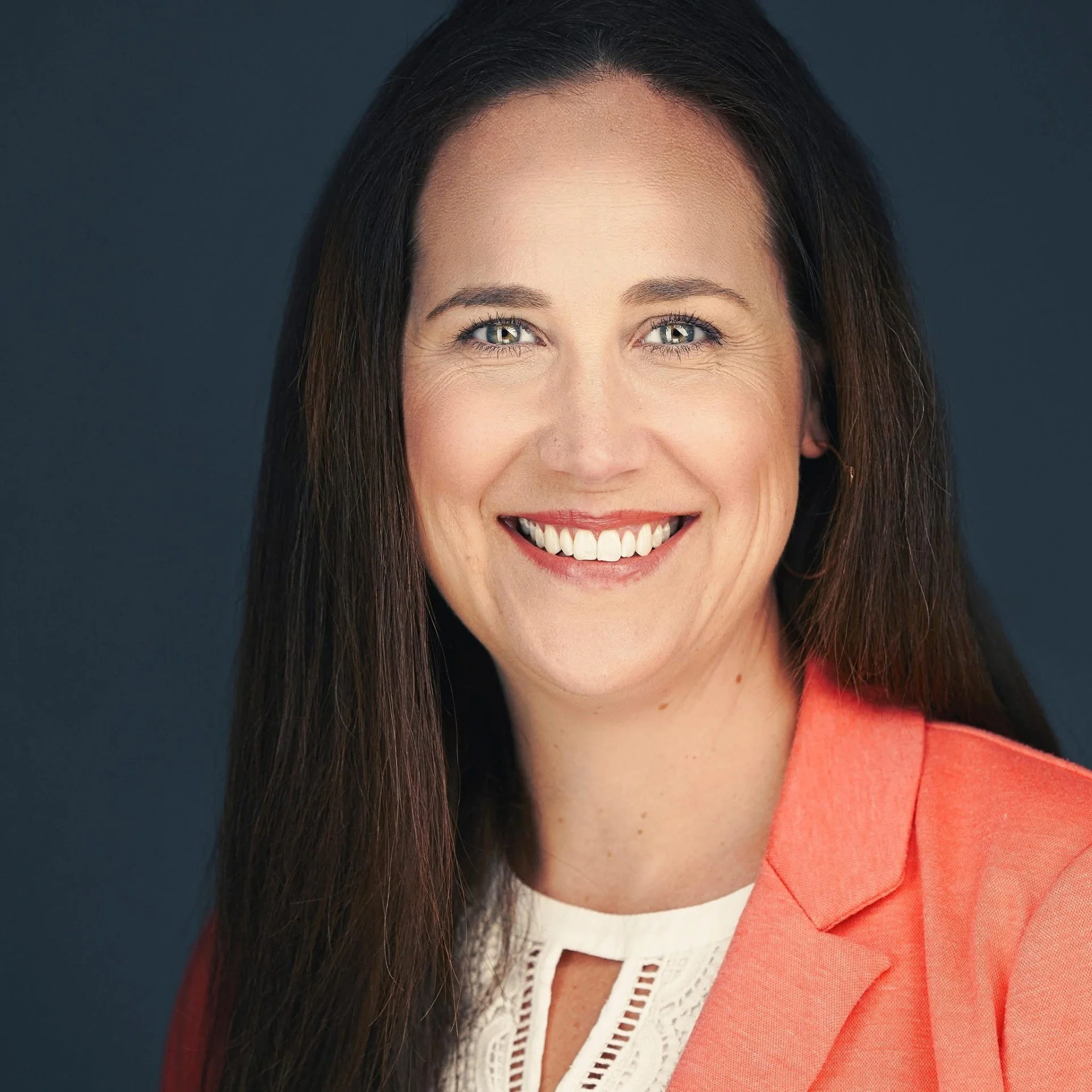A woman with long dark hair wearing a coral blazer and white top, smiling with teeth showing, against a dark background.