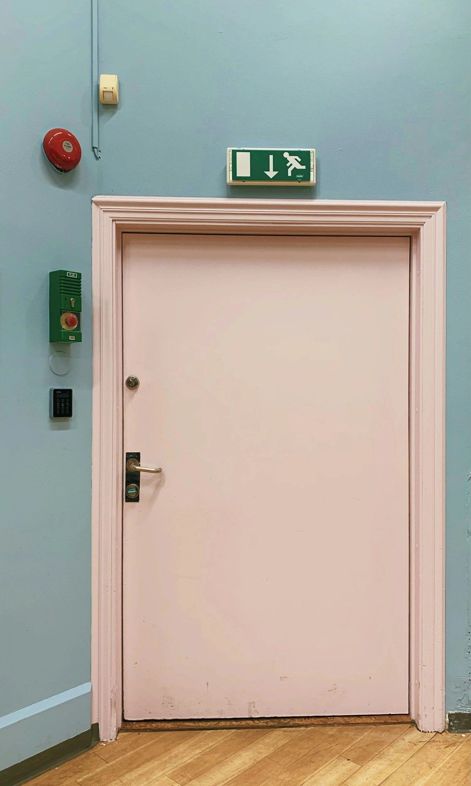 White door with a silver handle and a lock, set in a light pink door frame, against a light blue wall. Various security devices are mounted on the wall, including a green alarm button, a black keypad, and a red alarm bell. An emergency exit sign with a white running figure, a downward arrow, and a side arrow is mounted above the door. The floor is wooden.