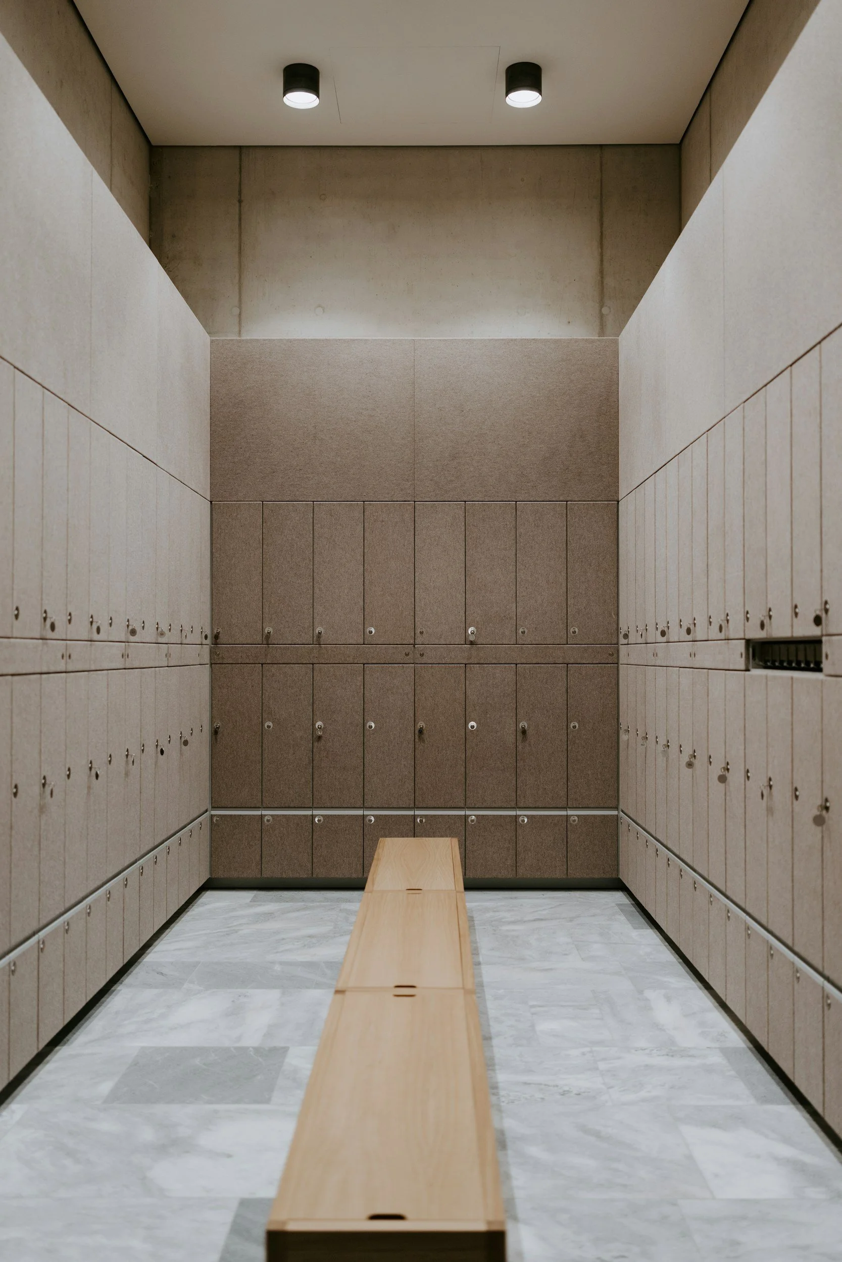 A locker room with beige lockers on both sides, a wooden bench in the center, and a plain ceiling with two recessed lights.