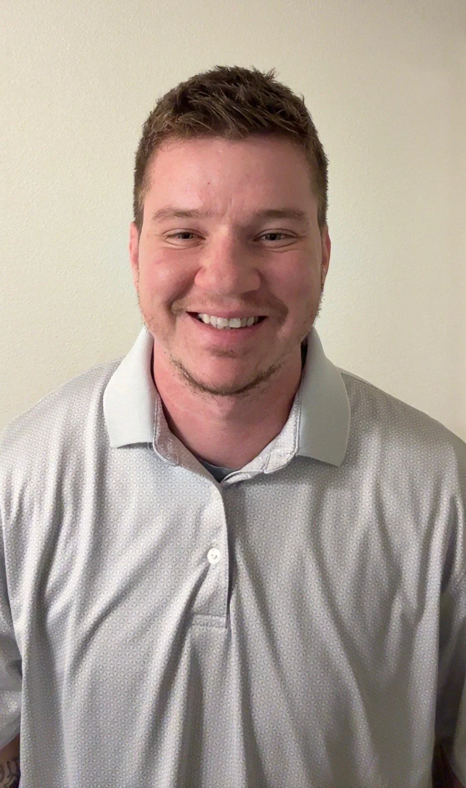Portrait of a smiling young man with short brown hair, wearing a light gray patterned collared shirt, standing against a plain beige wall.