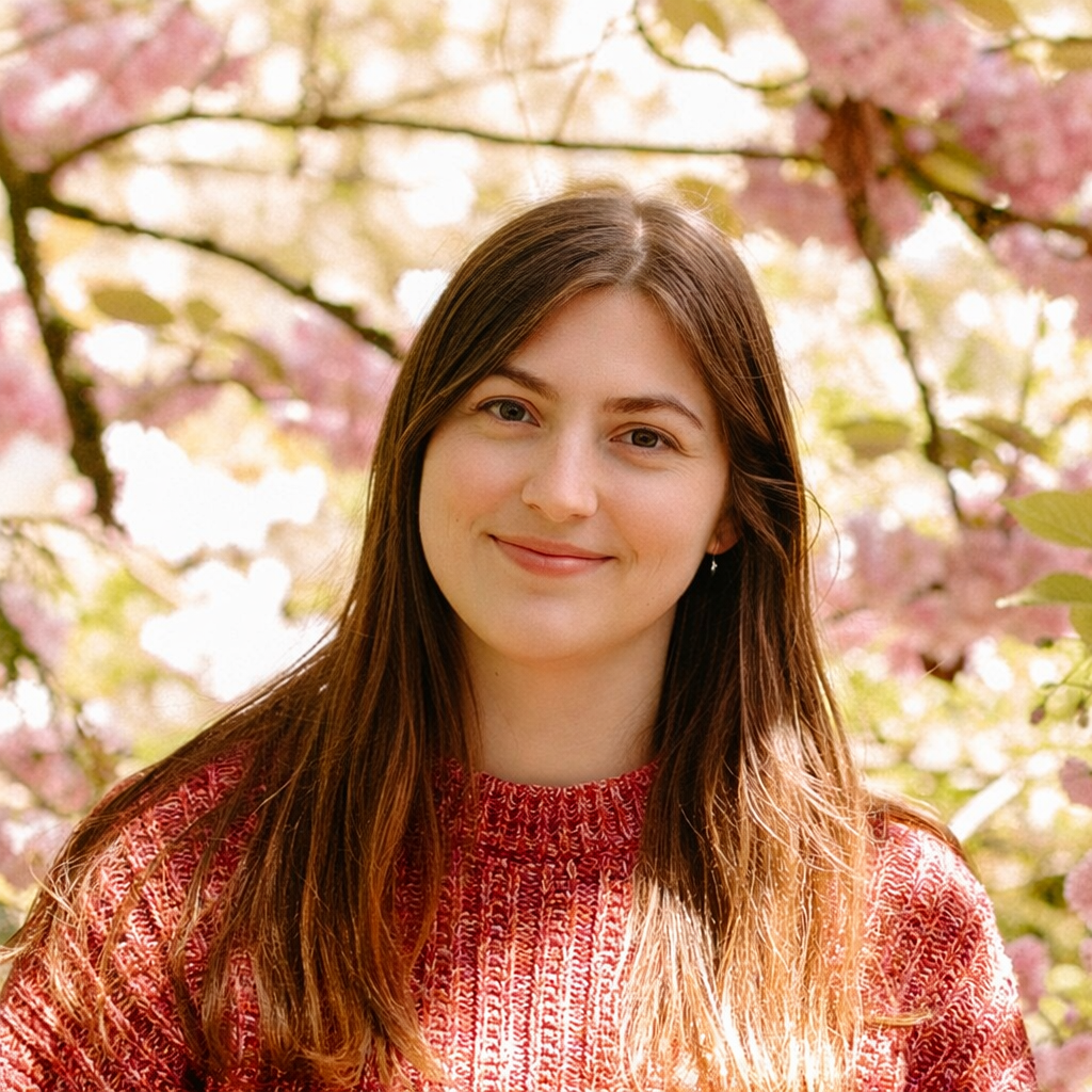 A young woman with long brown hair and a red patterned sweater standing outdoors among pink cherry blossoms.