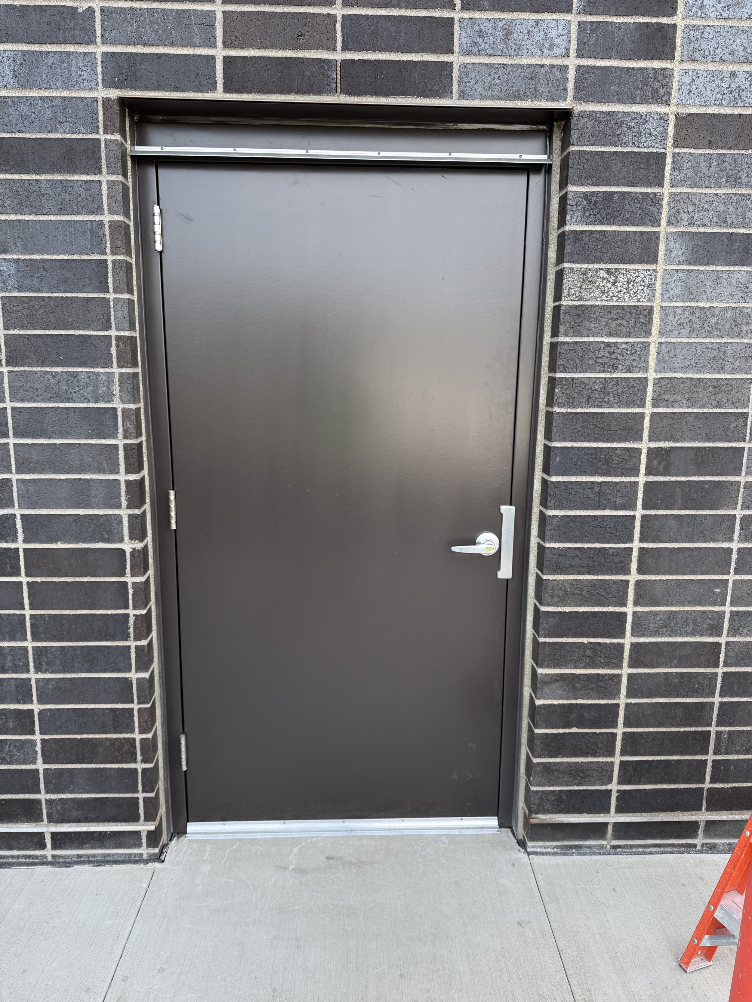 Plain black metal door with silver handle, set in a brick wall, with a small orange ladder visible at the bottom right.