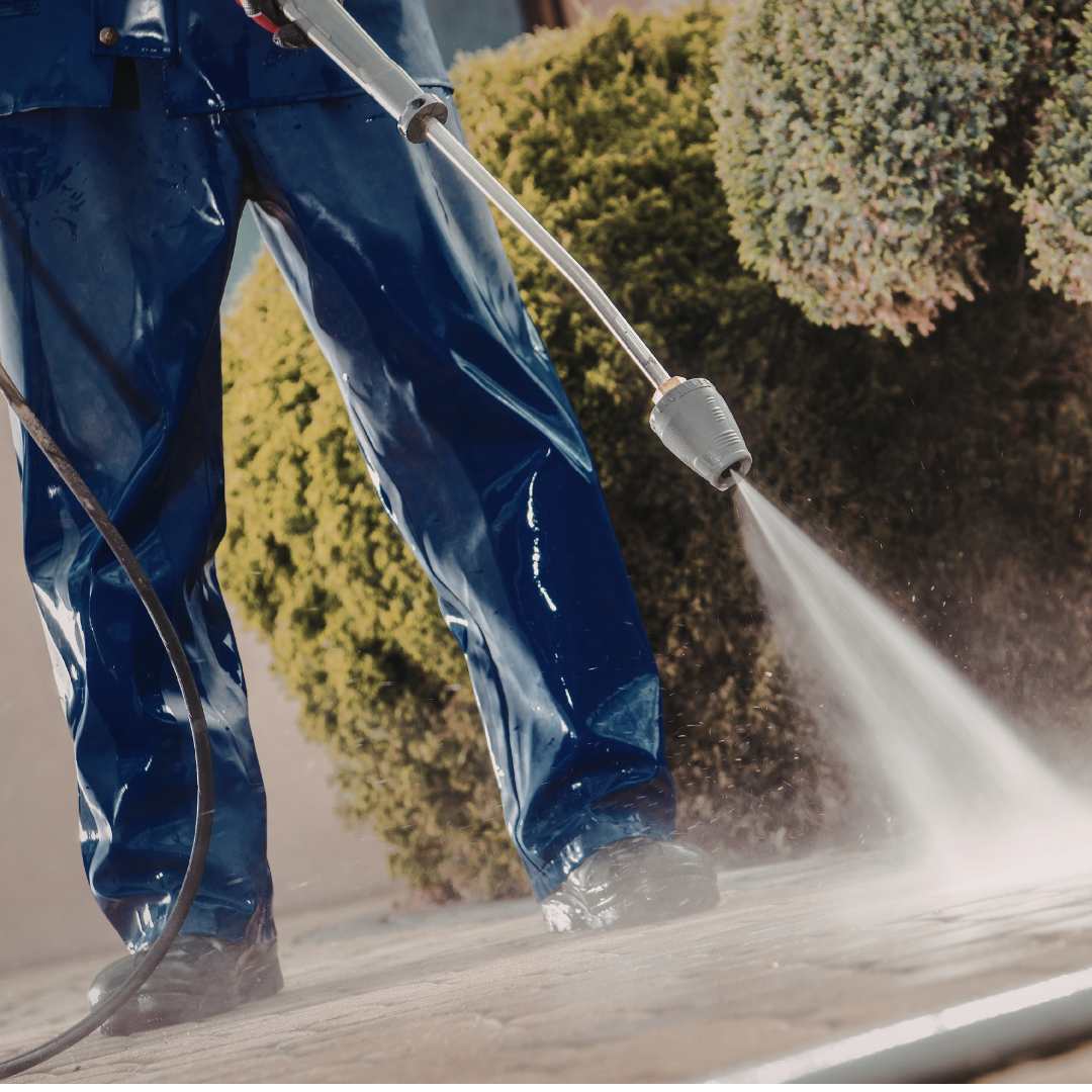 Person wearing blue waterproof suit, using a power washer to clean a surface outdoors with green bushes in the background.