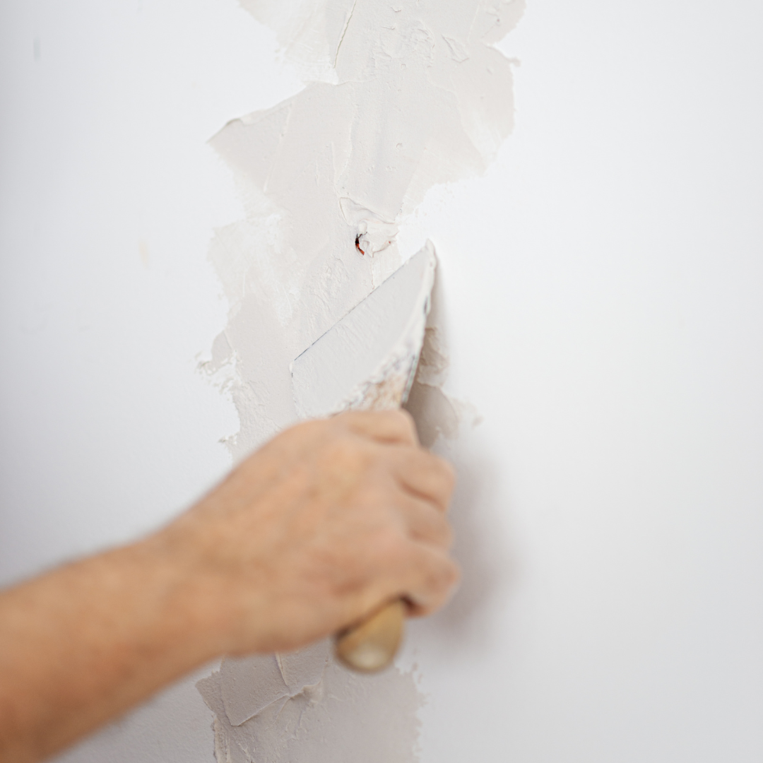 A person's hand is scraping peeling paint off a white wall with a putty knife.