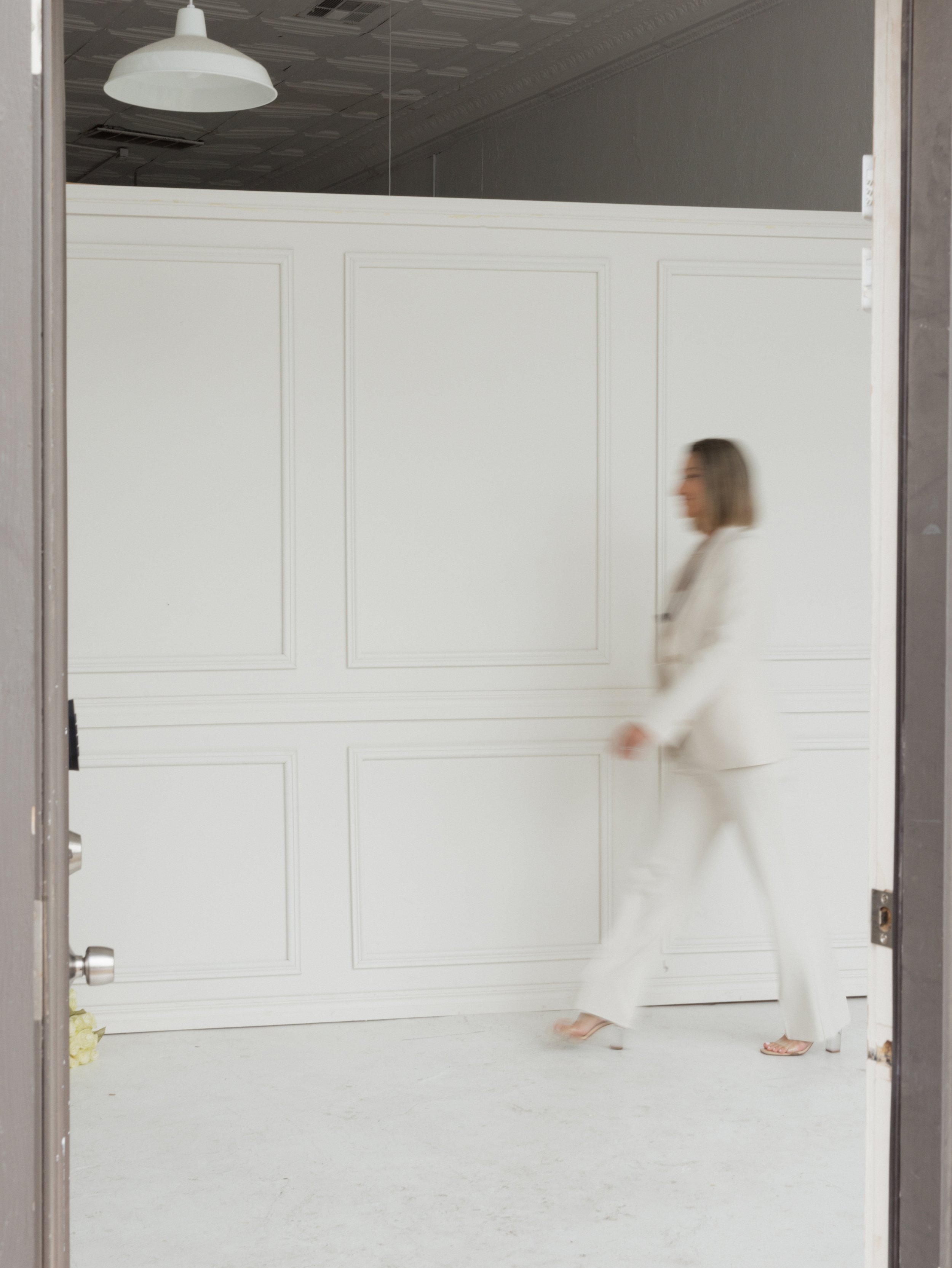 A woman dressed in a white outfit, walking past a white wall with paneling, seen through an open elevator door.
