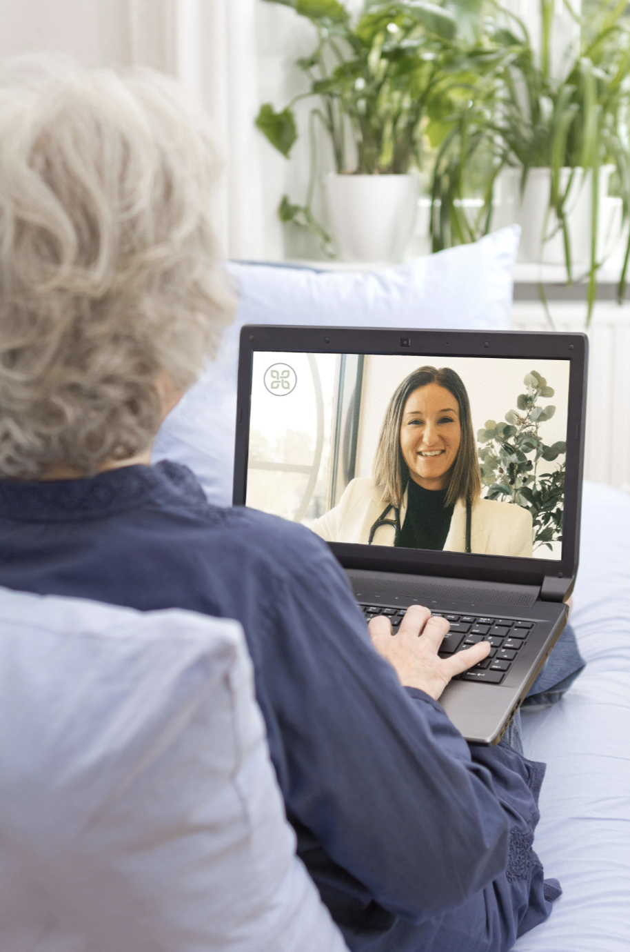 An elderly woman with white hair using a laptop for a video call with a woman in a white blazer, in a cozy room with green plants in the background.