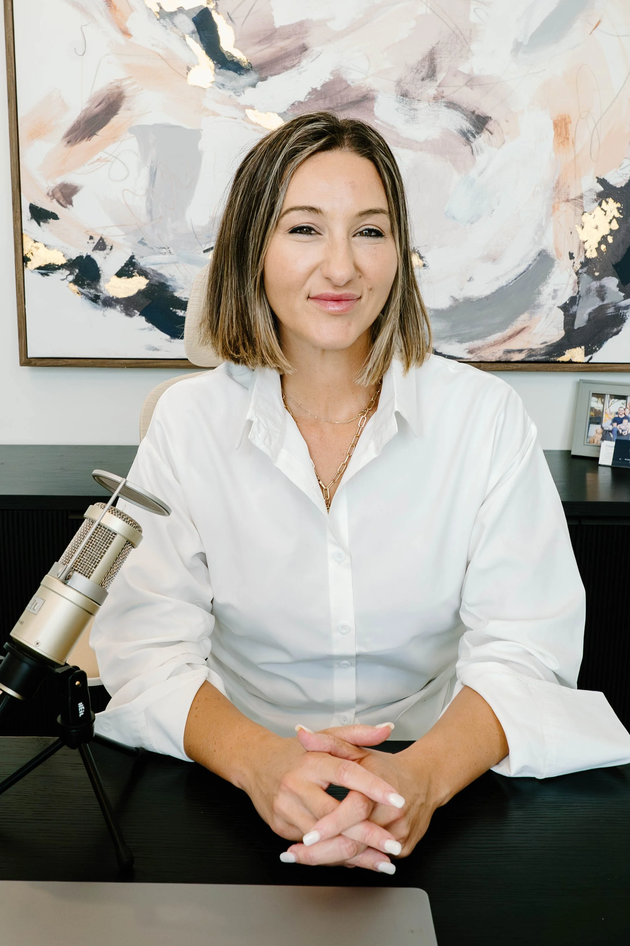 A woman with shoulder-length brown hair sitting at a desk in front of a microphone, wearing a white button-up shirt and jewelry, with a large abstract painting and framed photograph in the background.