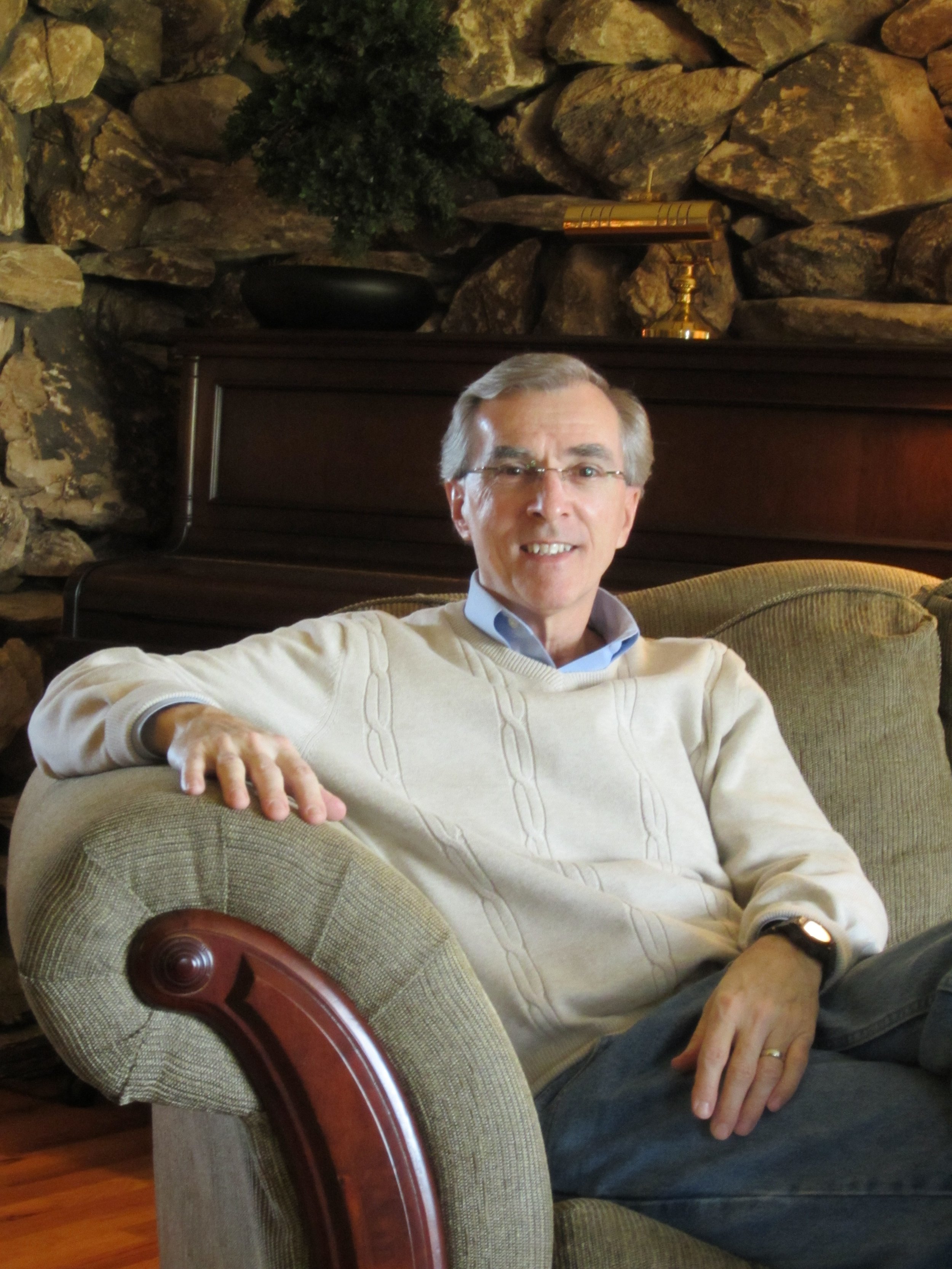 A middle-aged man with gray hair, glasses, wearing a cream-colored sweater, seated comfortably on a beige upholstered armchair in a cozy living room with stone walls and a wooden piano in the background.