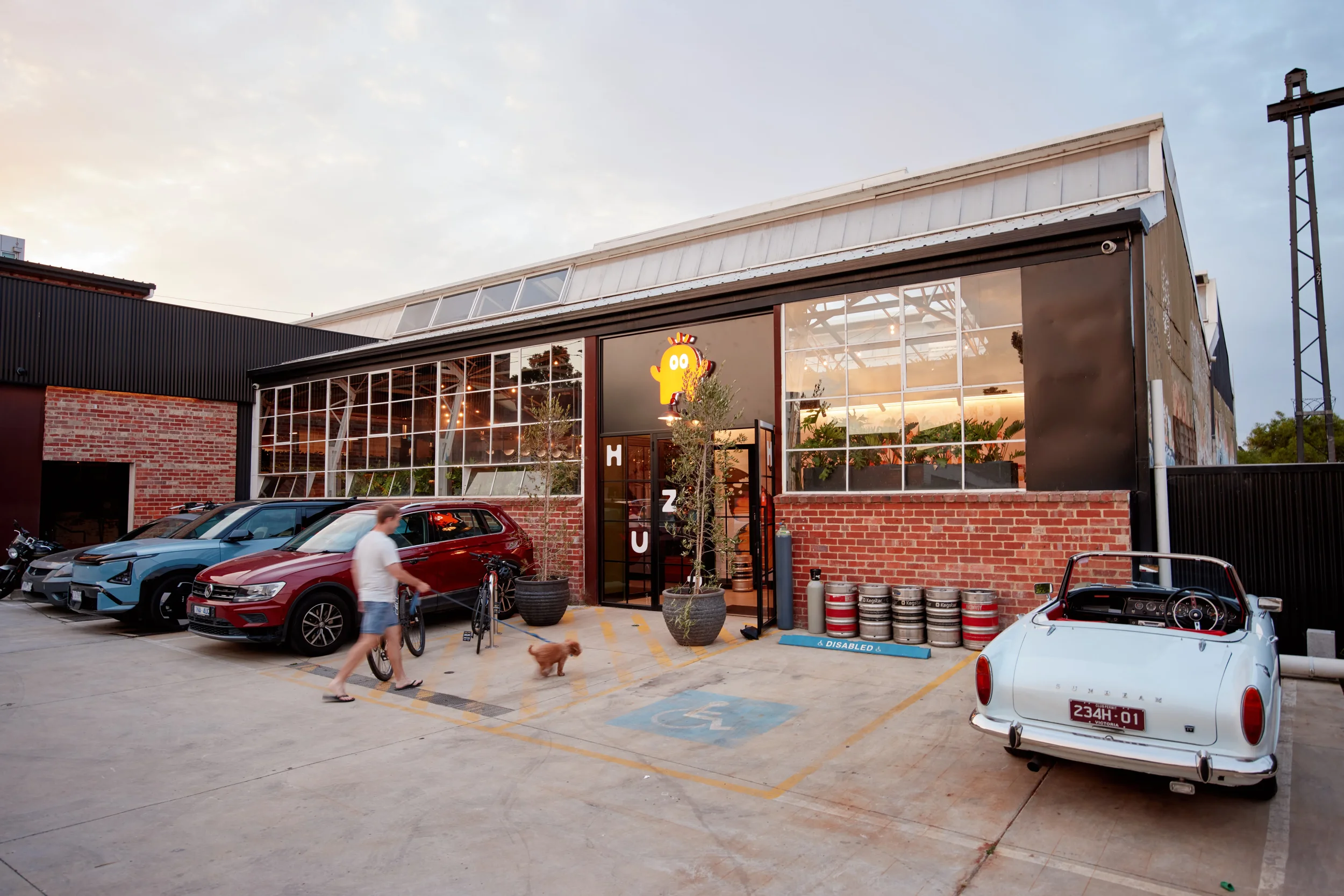 Exterior view of a modern industrial-style building with large glass windows and brick accents, parking lot with cars and a vintage convertible, man walking a dog, and bright sky at sunset.
