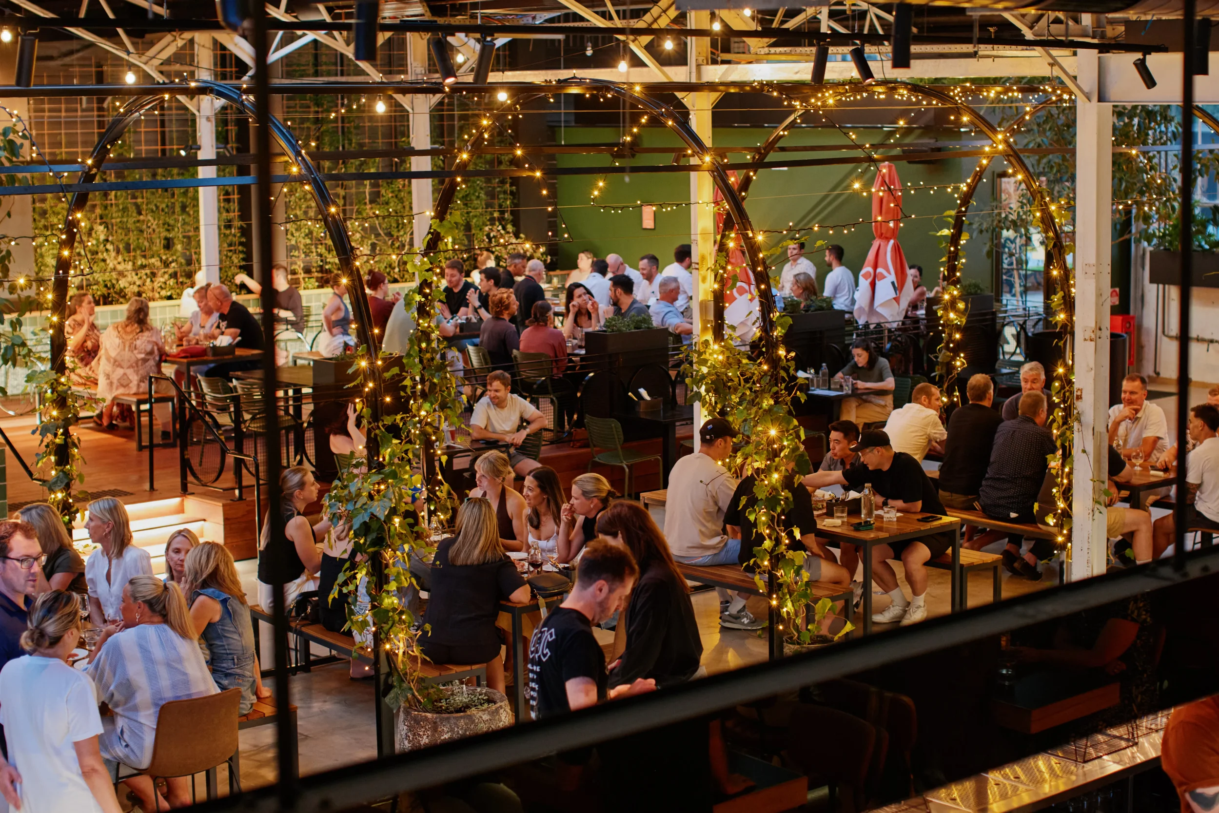 People dining at an outdoor restaurant or bar at night, decorated with string lights and greenery.