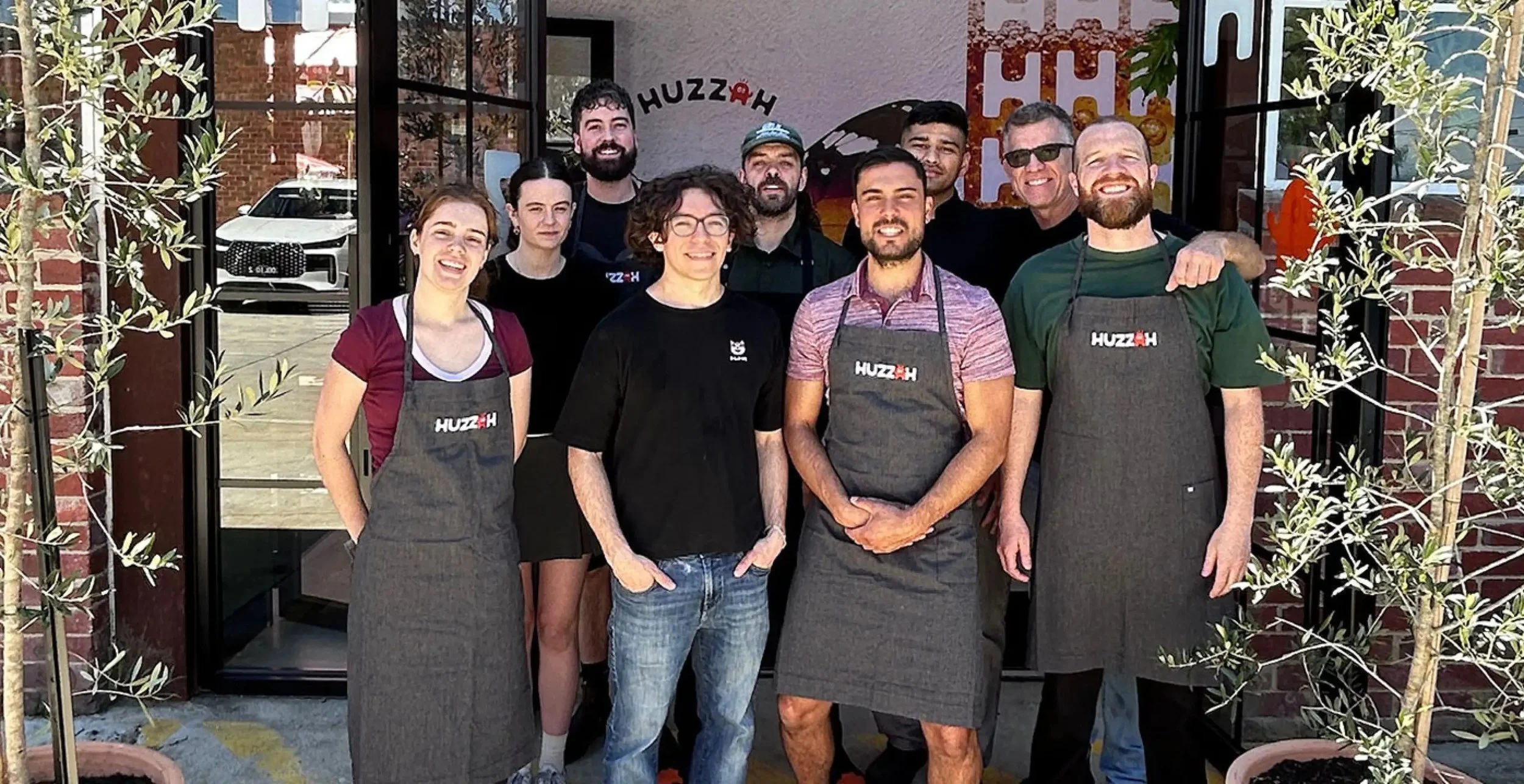Group of eleven people standing outside a restaurant or cafe, some wearing aprons with 'HUZZAH' logo, smiling at the camera, with a glass door, brick wall, and potted plants in the background.