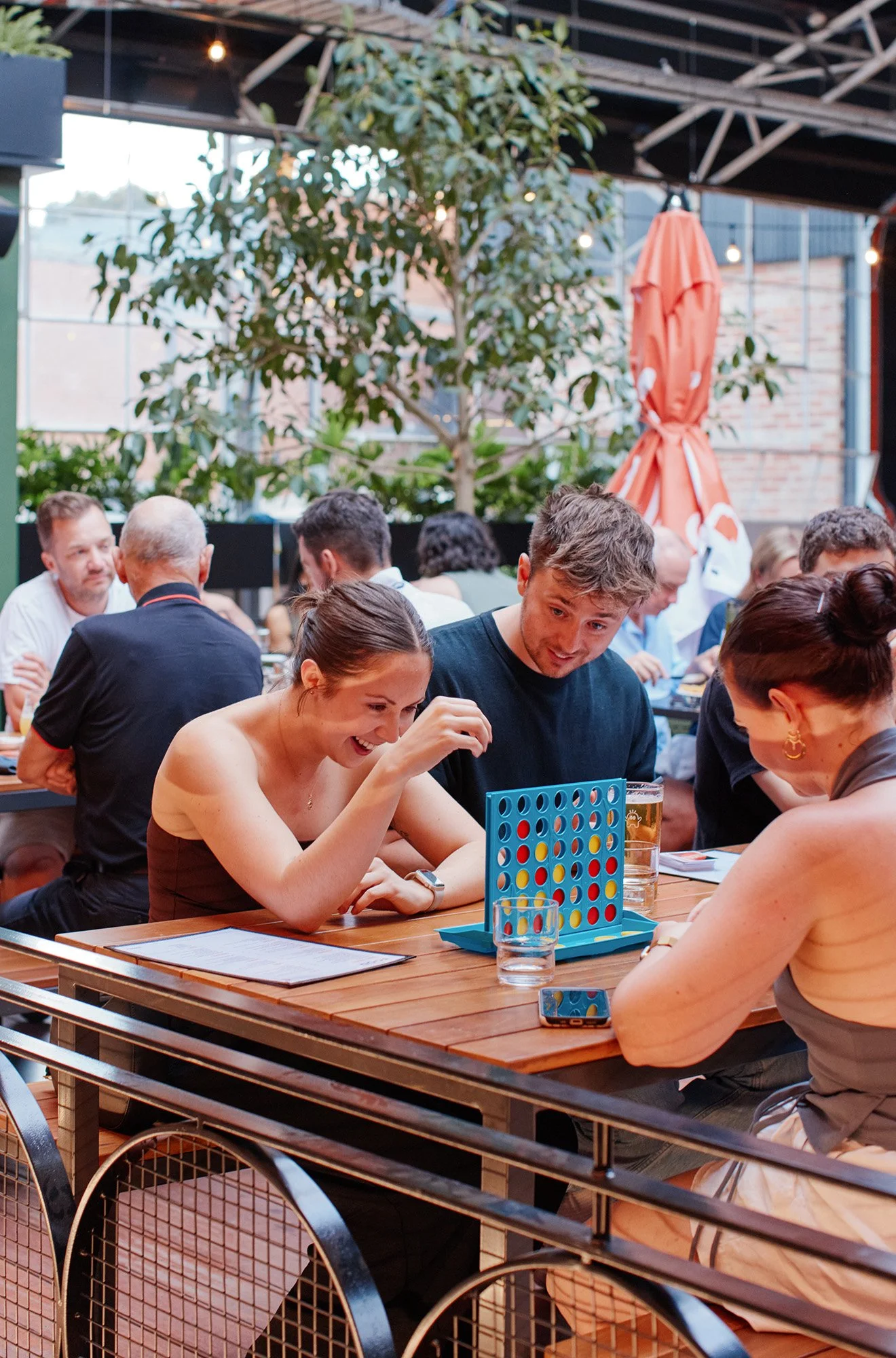 People playing Connect Four at a restaurant or cafe with outdoor seating, large green plant, and orange umbrella in the background.