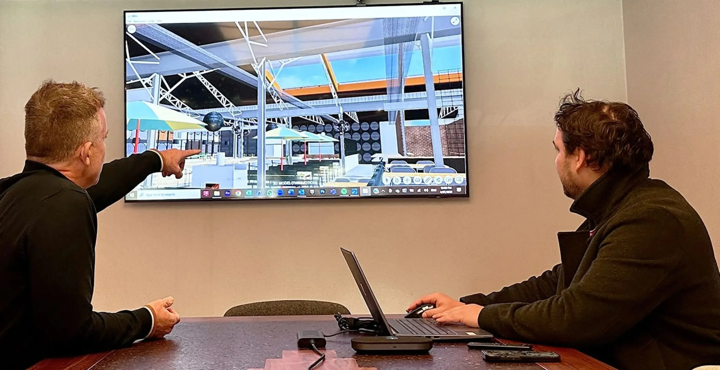 Two men discussing a 3D architectural model displayed on a wall-mounted screen in a meeting room.