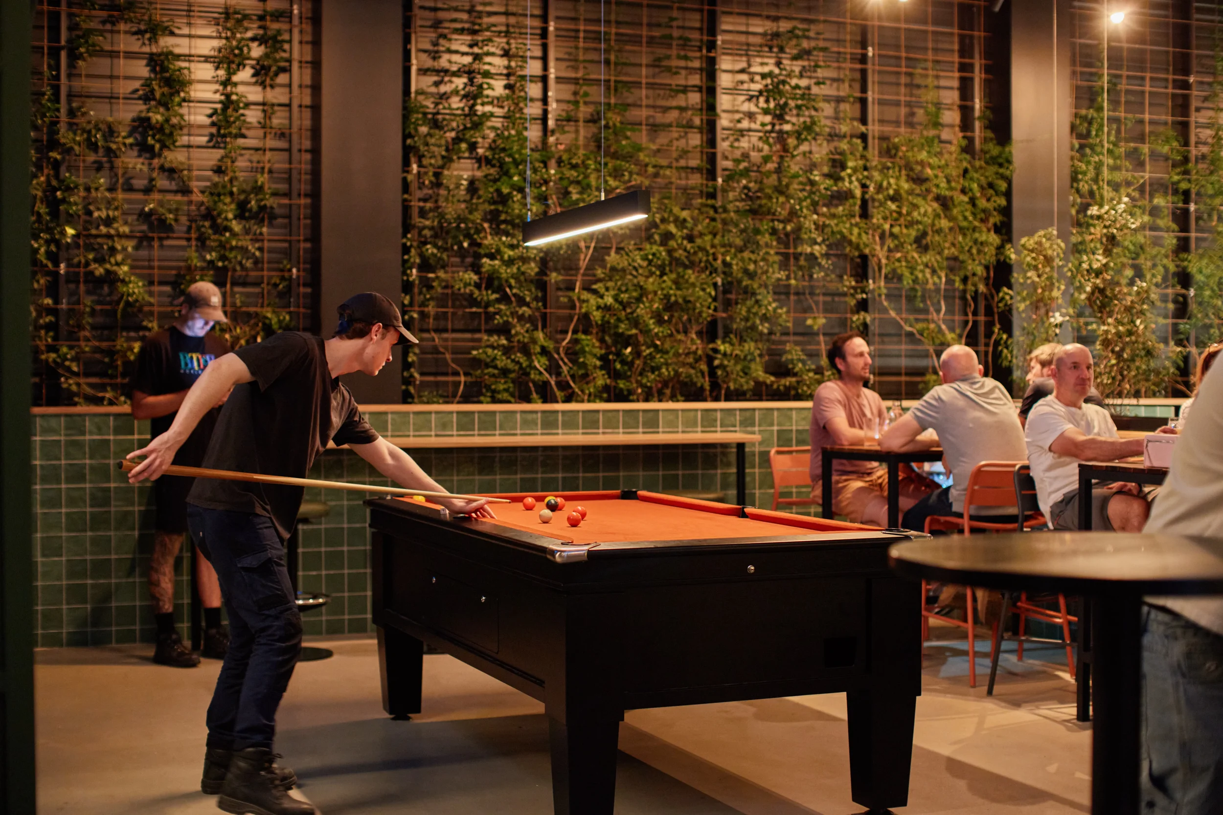 A young man wearing a black shirt and cap playing pool at a recreational venue while others sit at tables in the background in a cozy, dimly lit space with green tiled walls and large windows with plants outside.