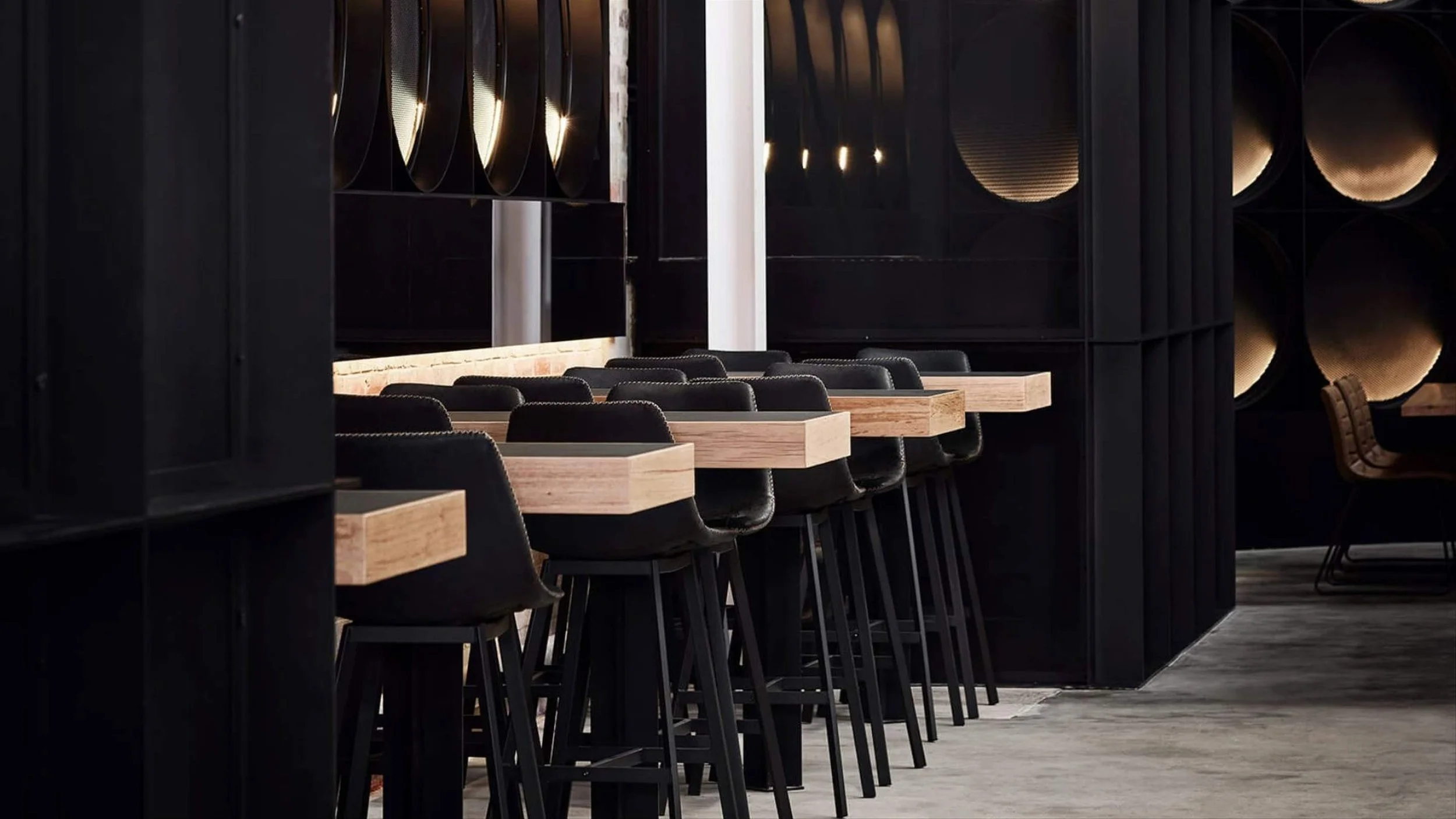 A modern bar or restaurant interior with a row of high black chairs and wooden bar tables against a dark wall with circular wall decorations.