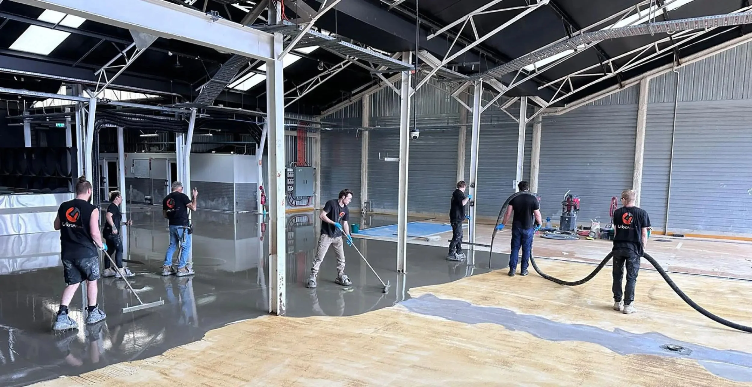 Workers applying a concrete overlay on a large indoor floor using various tools, with some spreading and smoothing the concrete surface.