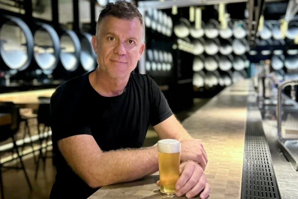 A man sitting at a bar holding a glass of beer, with rows of beer taps and shelves behind him.