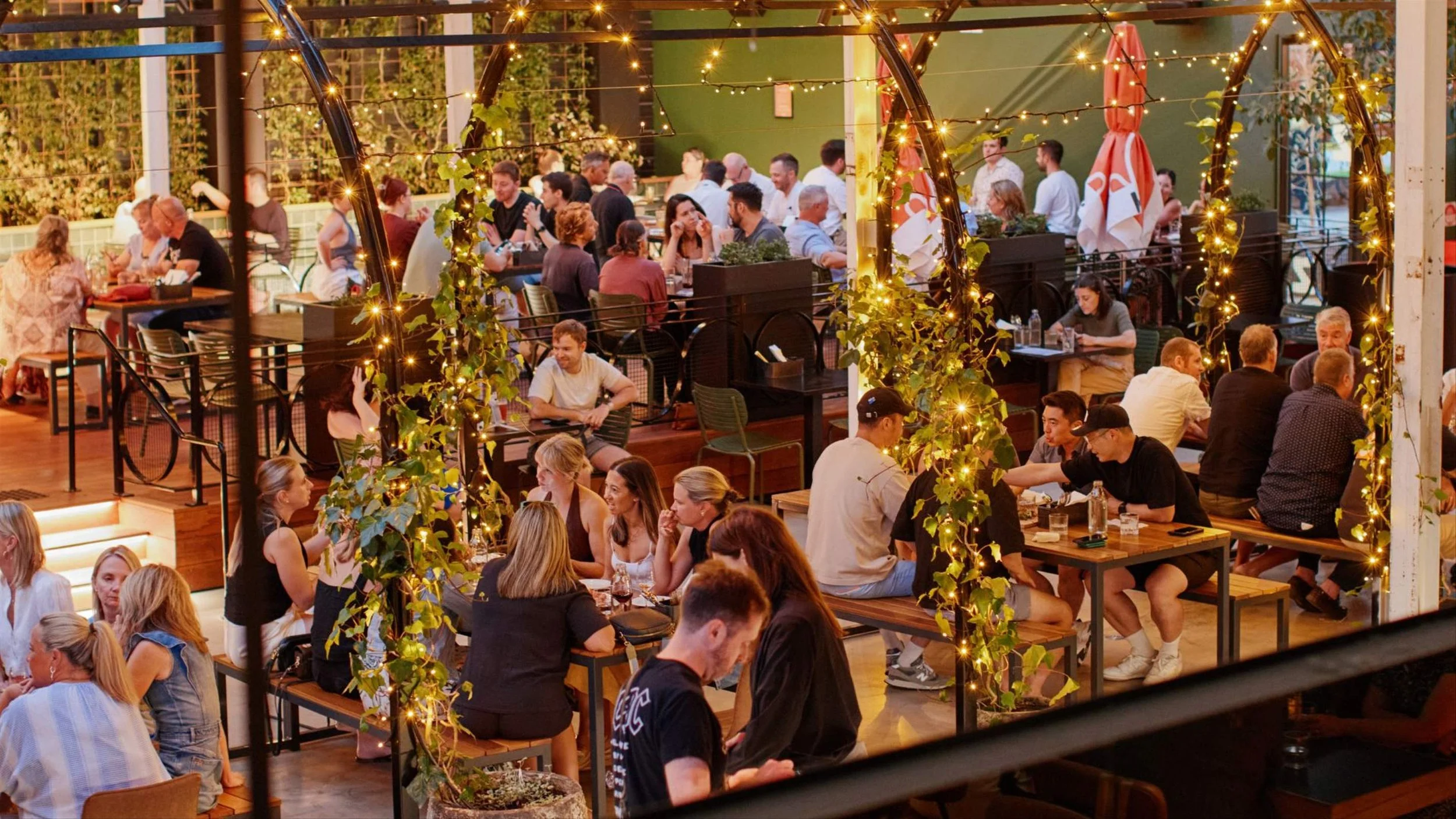 People dining in an indoor/outdoor restaurant decorated with string lights and greenery during the evening.