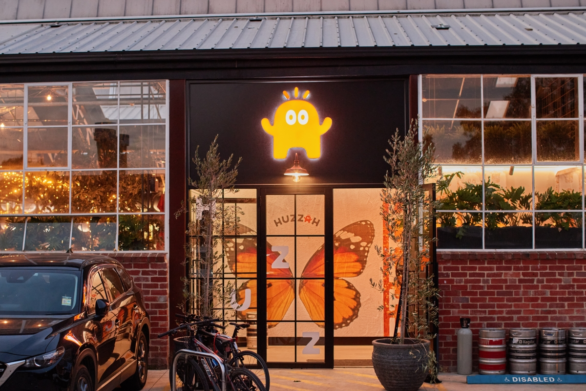 Exterior of a building with a neon yellow cartoon ghost sign above the entrance. The door has a butterfly mural behind it. Two bicycles and a parked black car are in front of the building. Glass windows reveal indoor plants and warm lighting.