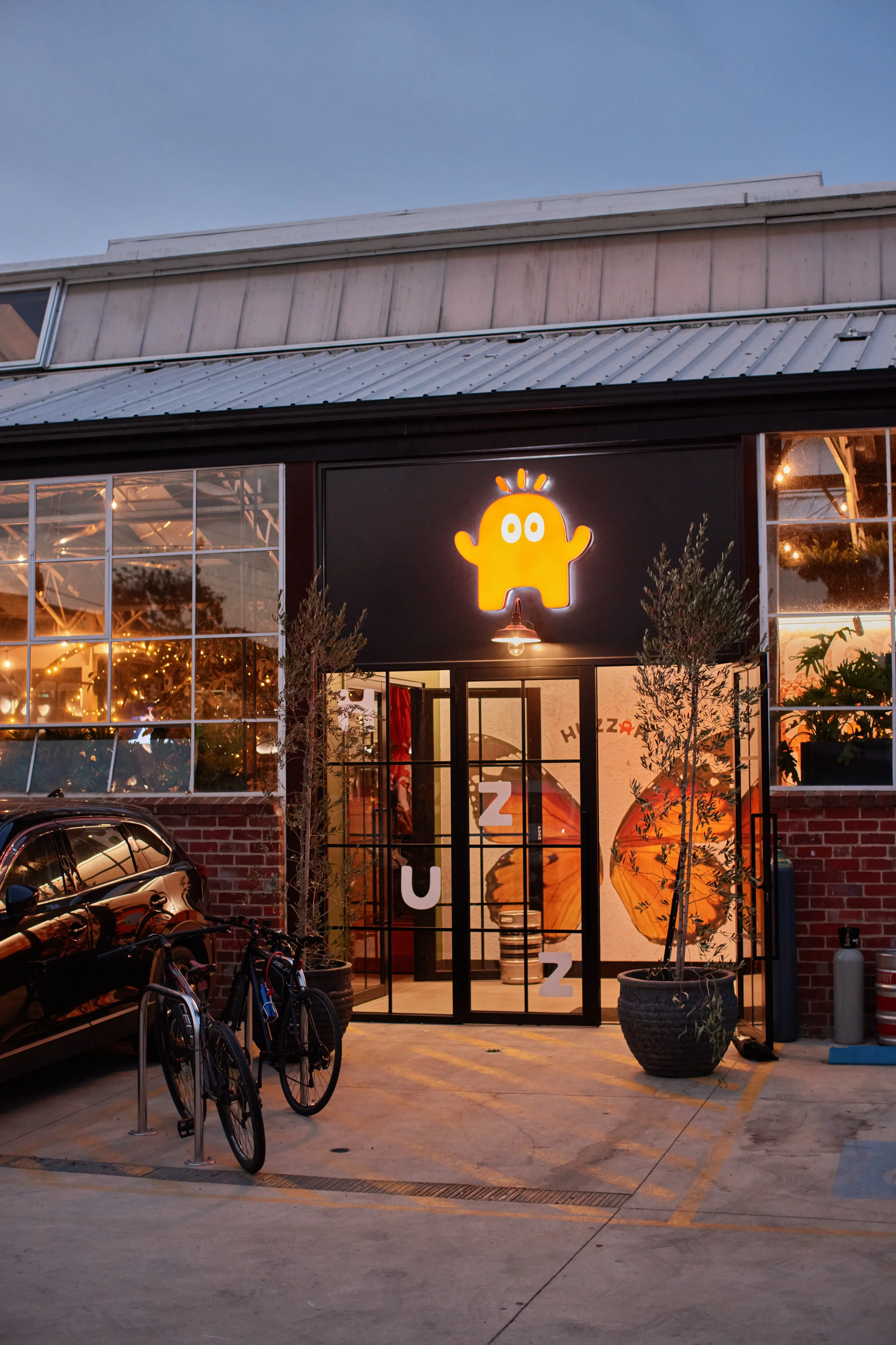 Outdoor view of a building with a black facade, decorated with a yellow neon mascot on top. The entrance has glass doors and is illuminated from inside. Two potted trees are positioned on either side of the entrance, with bicycles and a parked car nearby.