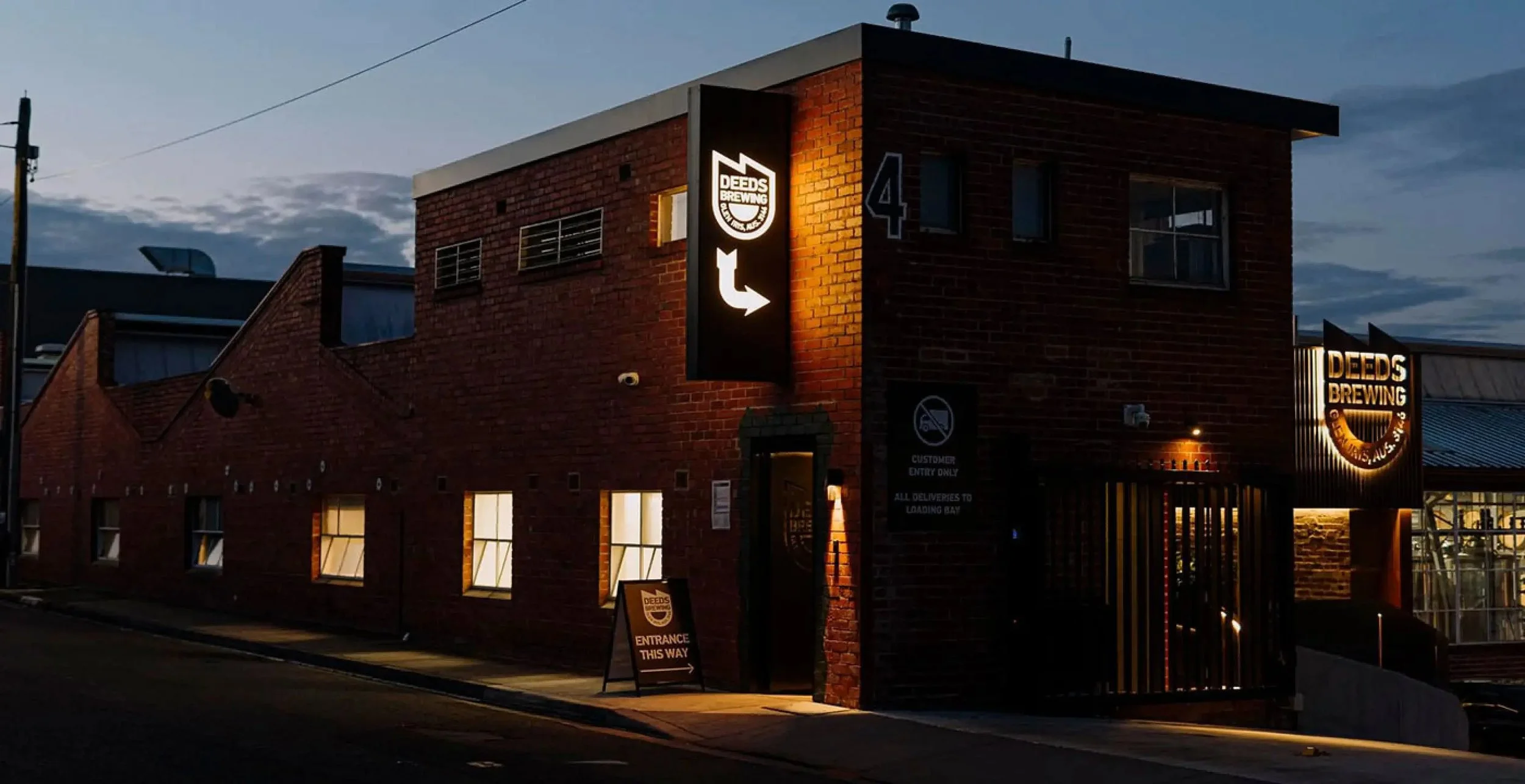 Night view of a brick building with illuminated sign for Deeds Brewing and an arrow pointing left. There are several windows with light inside, and a sidewalk with a sign indicating entrance. The sky is dark with some clouds.