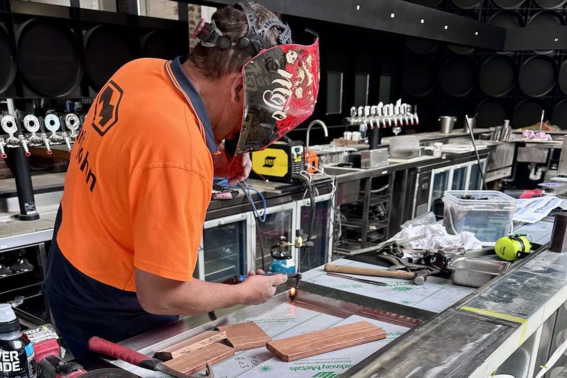 A man welding pieces of wood in a workshop or brewery with various tools, equipment, and containers on the work surface.