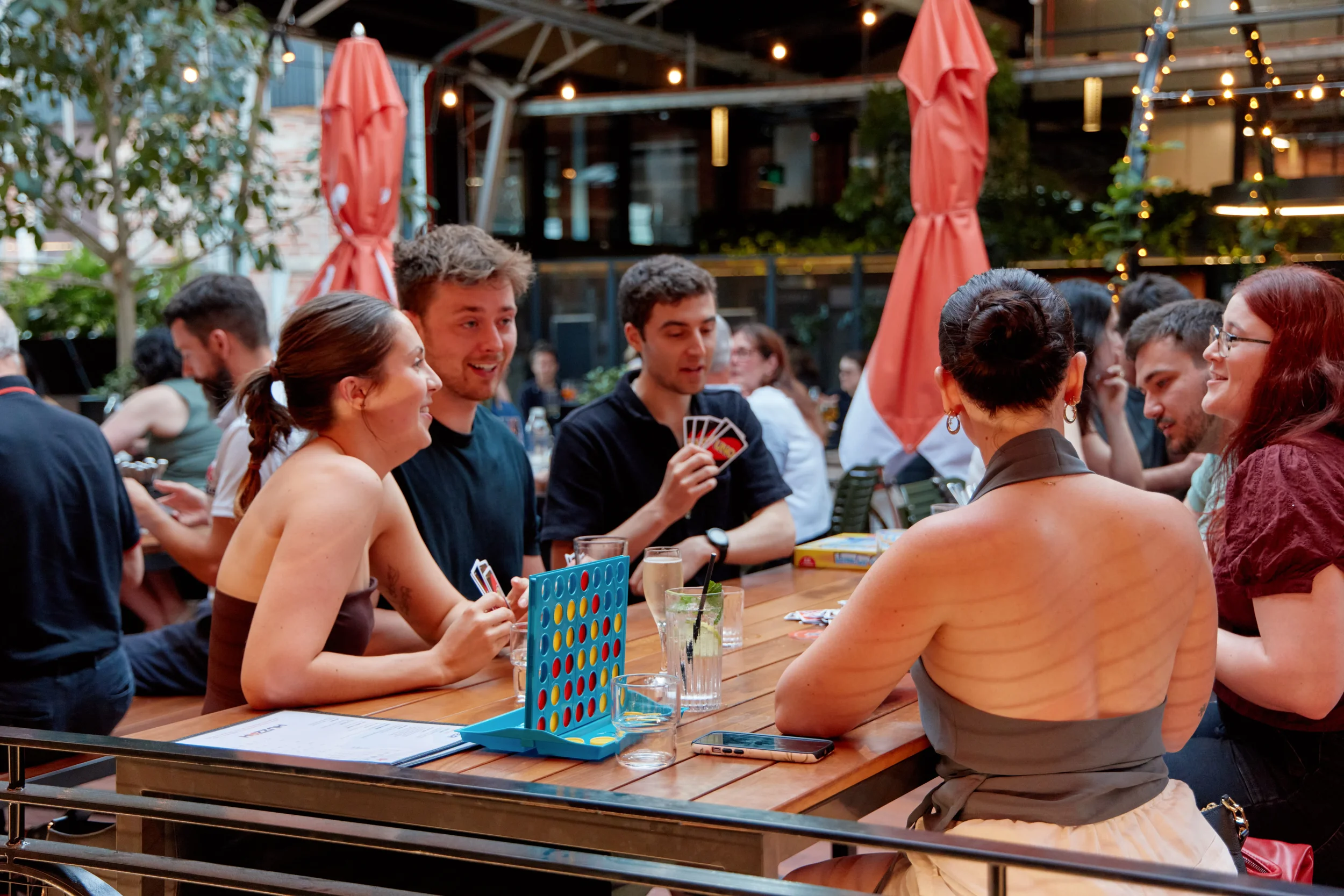 People playing Connect Four and chatting at an outdoor bar or patio with string lights and umbrellas.