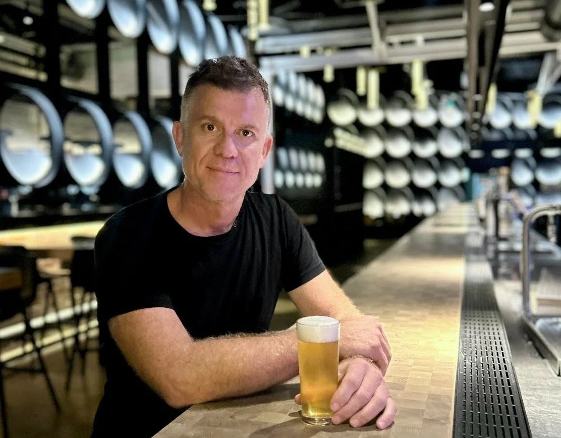 A man sitting at a bar holding a glass of beer, with a background of shelves filled with circular kegs.