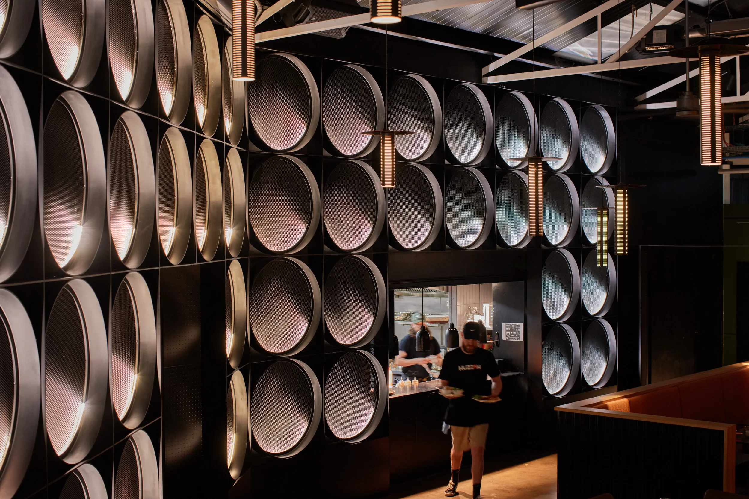 A modern restaurant interior featuring a wall decorated with large, rounded, metallic speakers or art pieces arranged in a grid pattern. There are hanging pendant lights with vertical light strips illuminating the space. In the background, a window into the kitchen shows staff preparing food, with a man in a black shirt and shorts walking past, holding plates.
