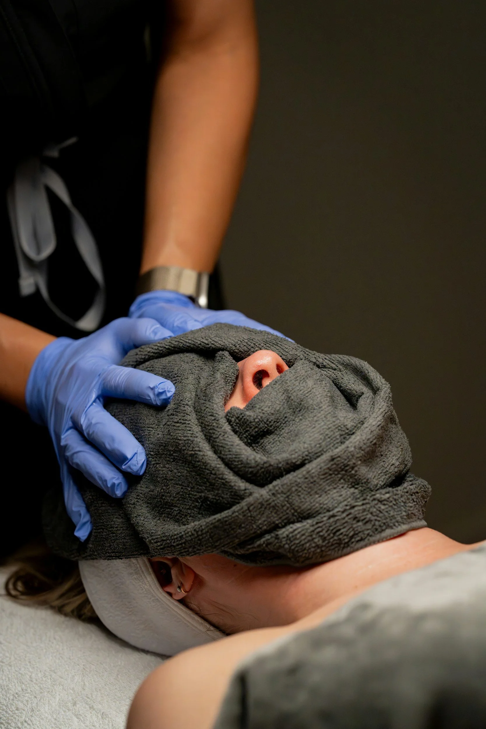 Person receiving facial treatment with towel wrapped around face, lying on a spa table, wearing a headband, and healthcare worker wearing blue gloves, black uniform, and watch.