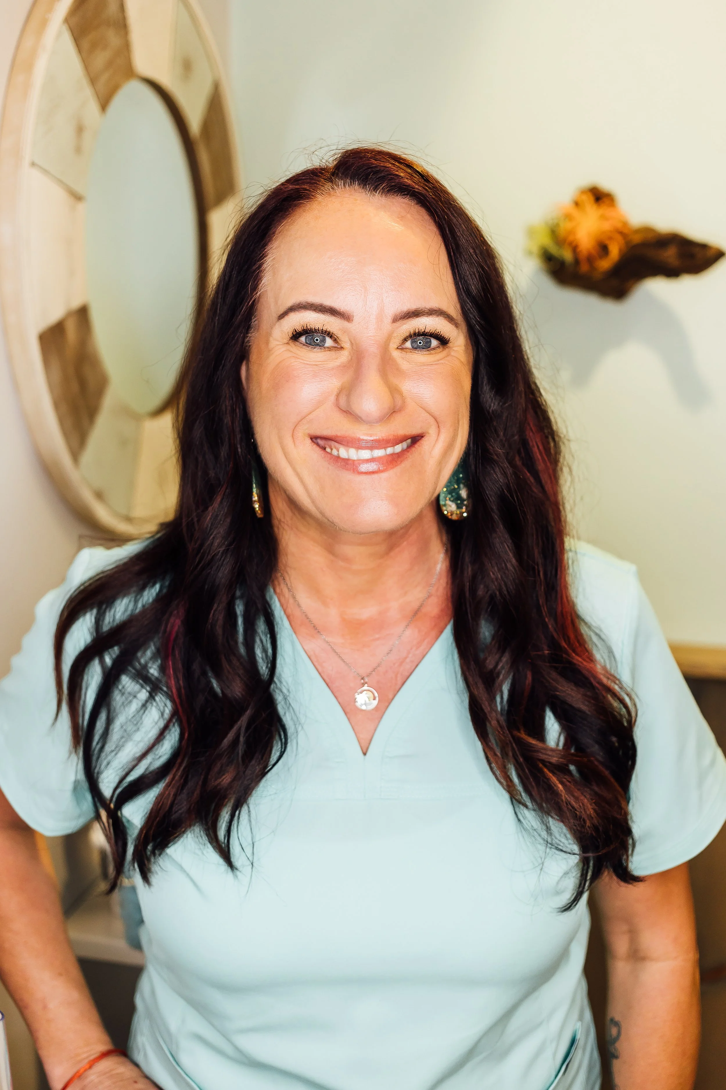 A smiling woman with long dark hair wearing scrubs, standing indoors.