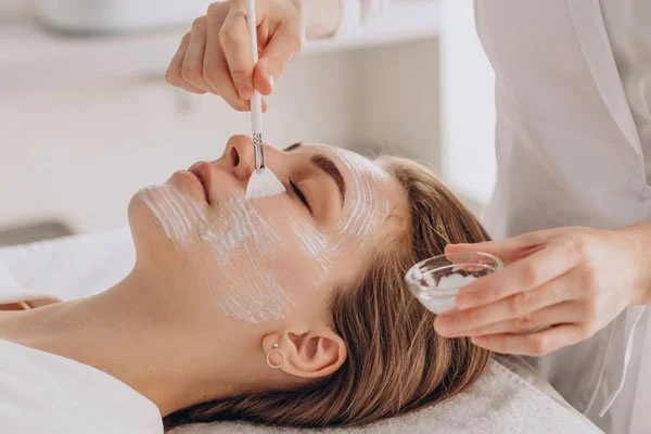 Esthetician applying facial mask to woman lying on treatment bed