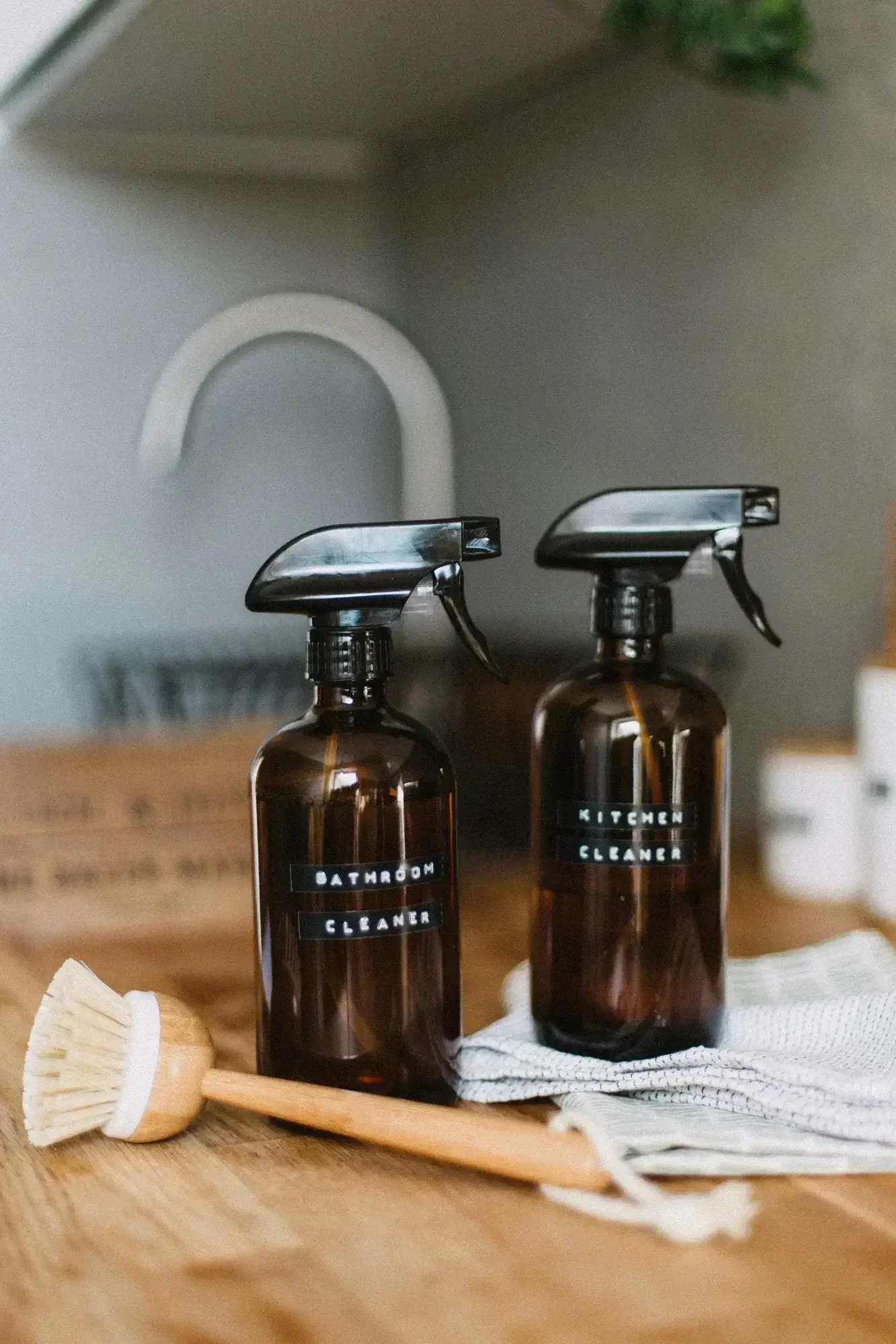 two dark brown generic but stylish cleaner bottles sitting on a wooden countertop near a rag, and by a sink