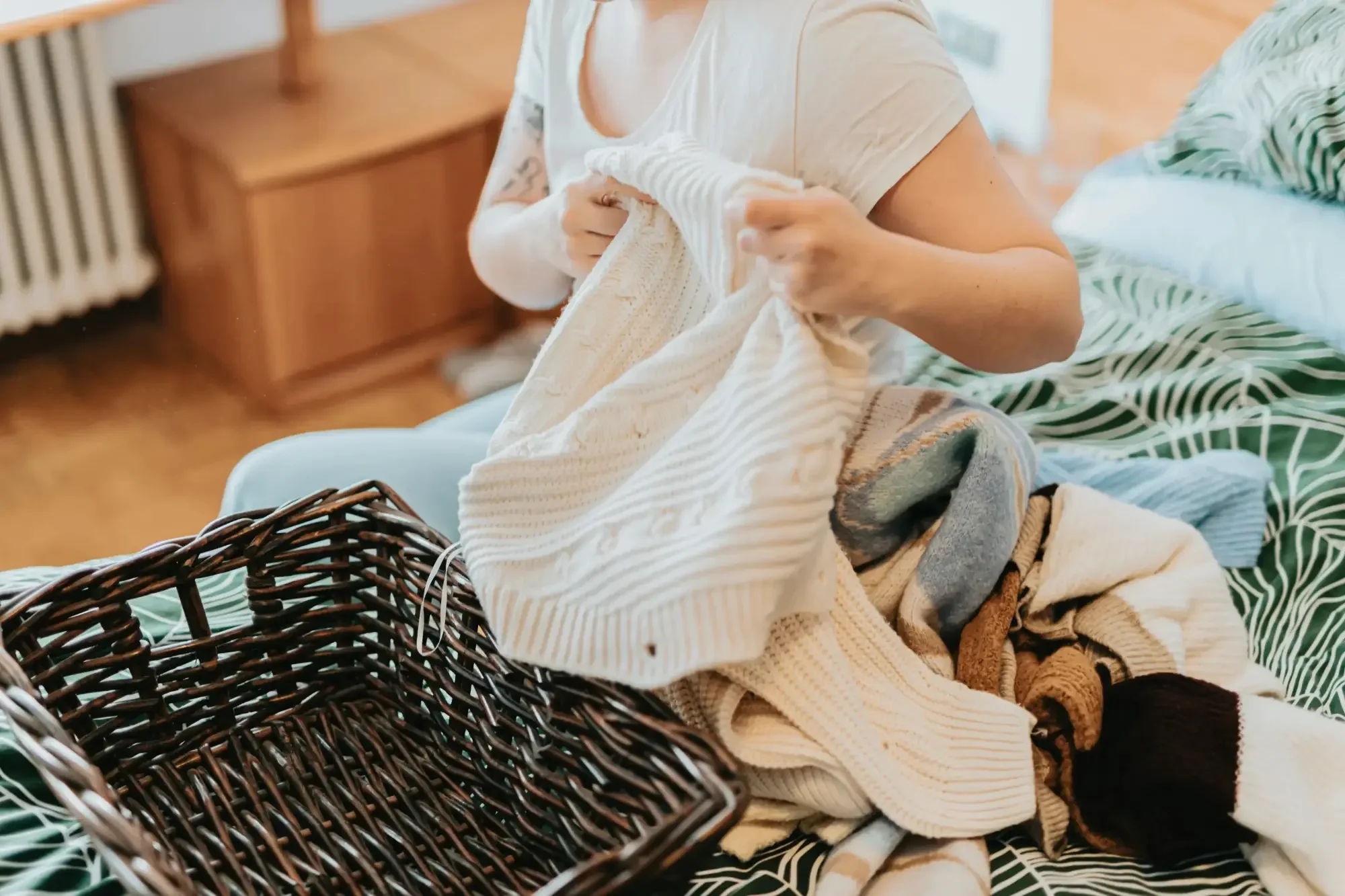 a woman sitting on a bed next to an empty basket, folding clothes & blankets to put away