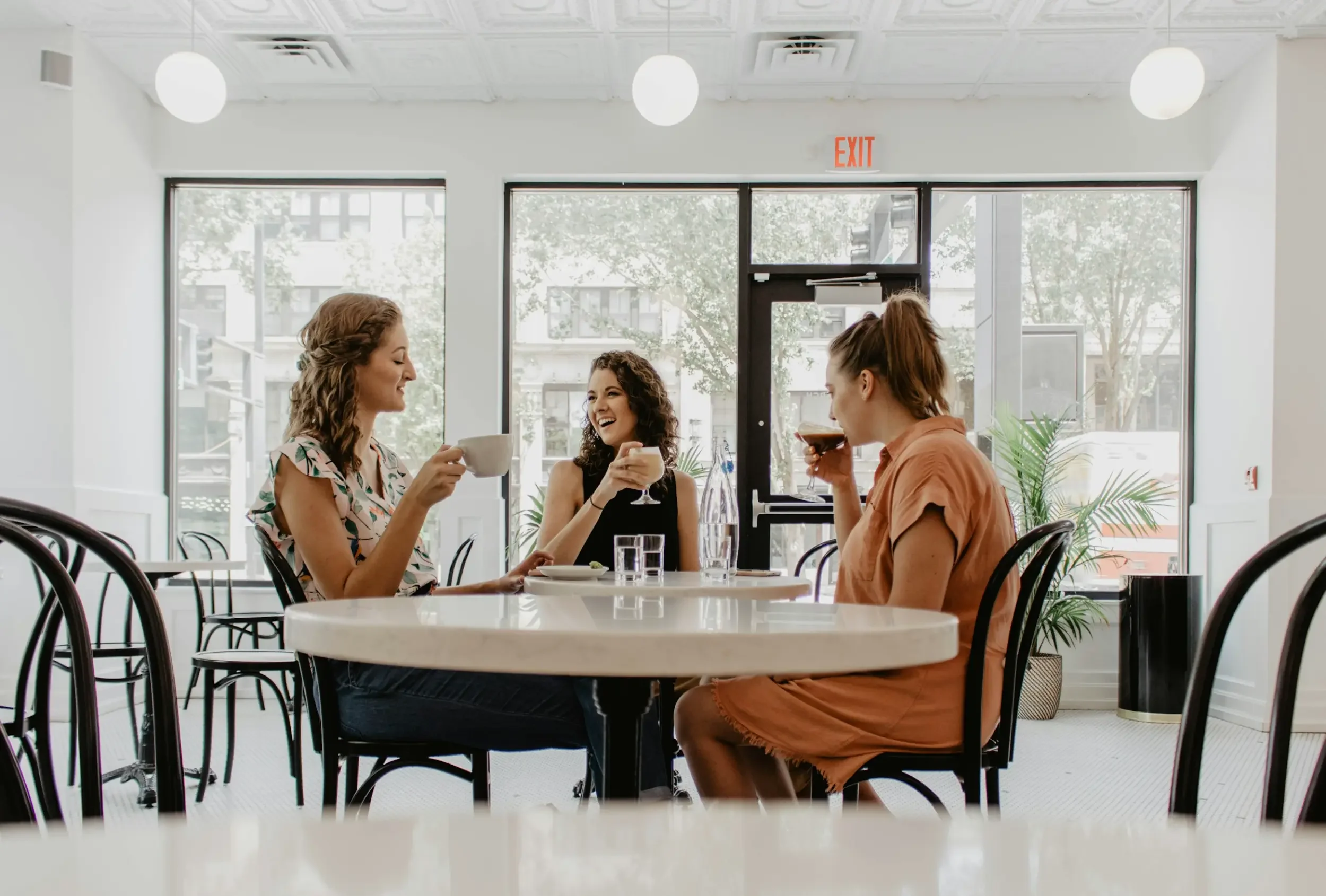 Three women laugh and enjoy coffee together in a bright, modern café—perfect for relaxing in stylish commercial common areas.