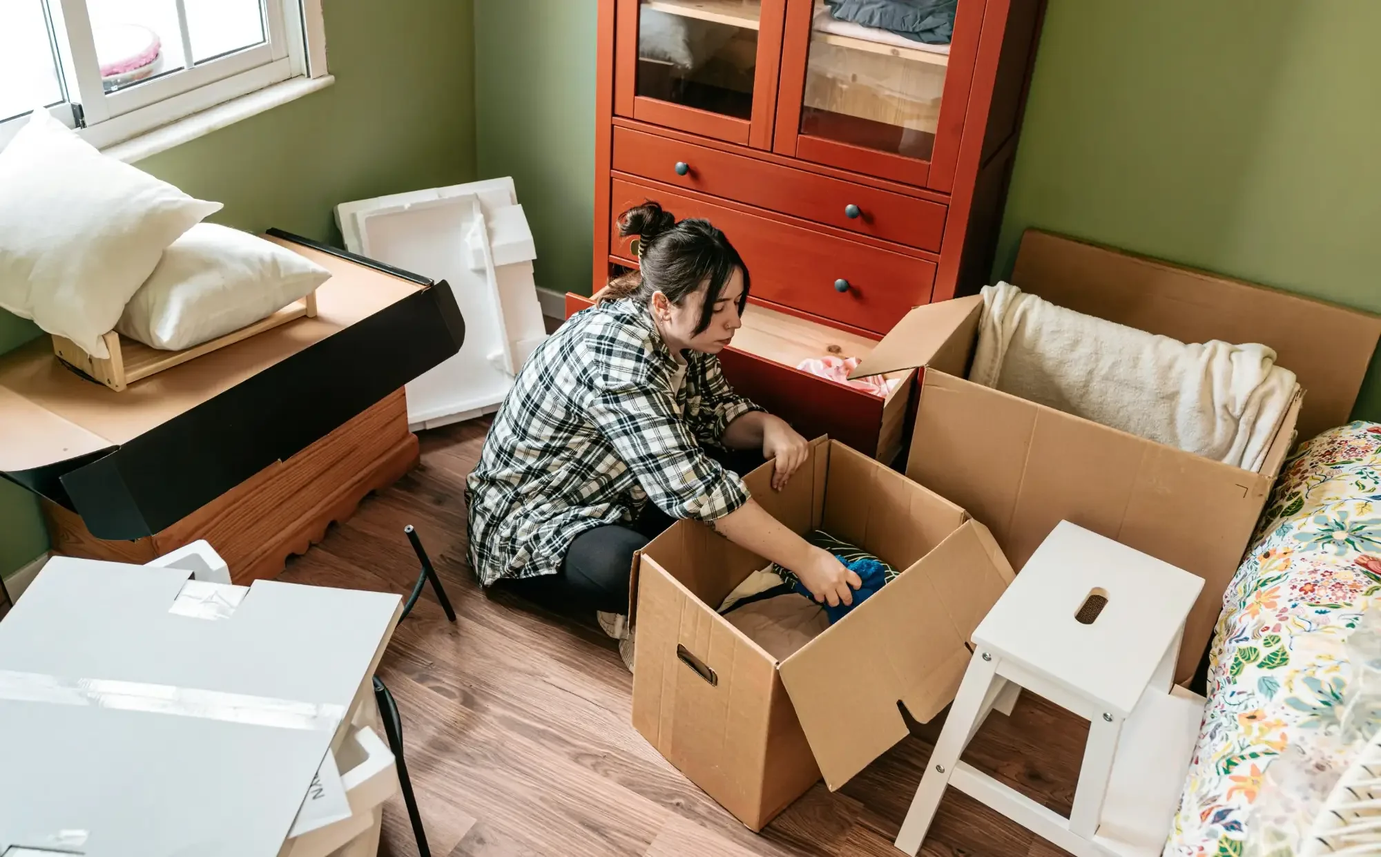 A woman sits on the floor packing clothes into a box, preparing for a move out clean among unpacked furniture and moving supplies.
