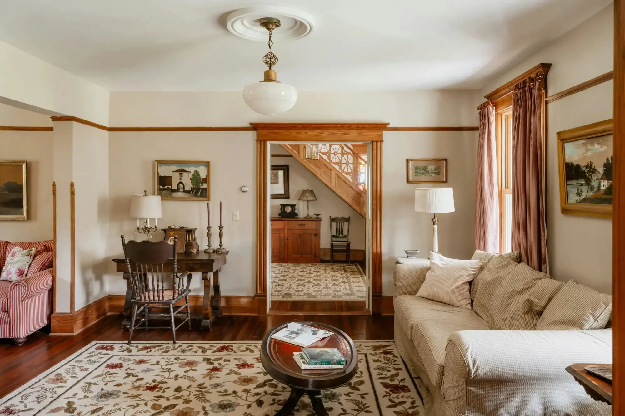 view of living area and entryway stairwell, bright light coming through the windows, dark hardwood floors & carpets, white & beige painted walls with wooden trim and antique-themed furnishings alongside modern couches