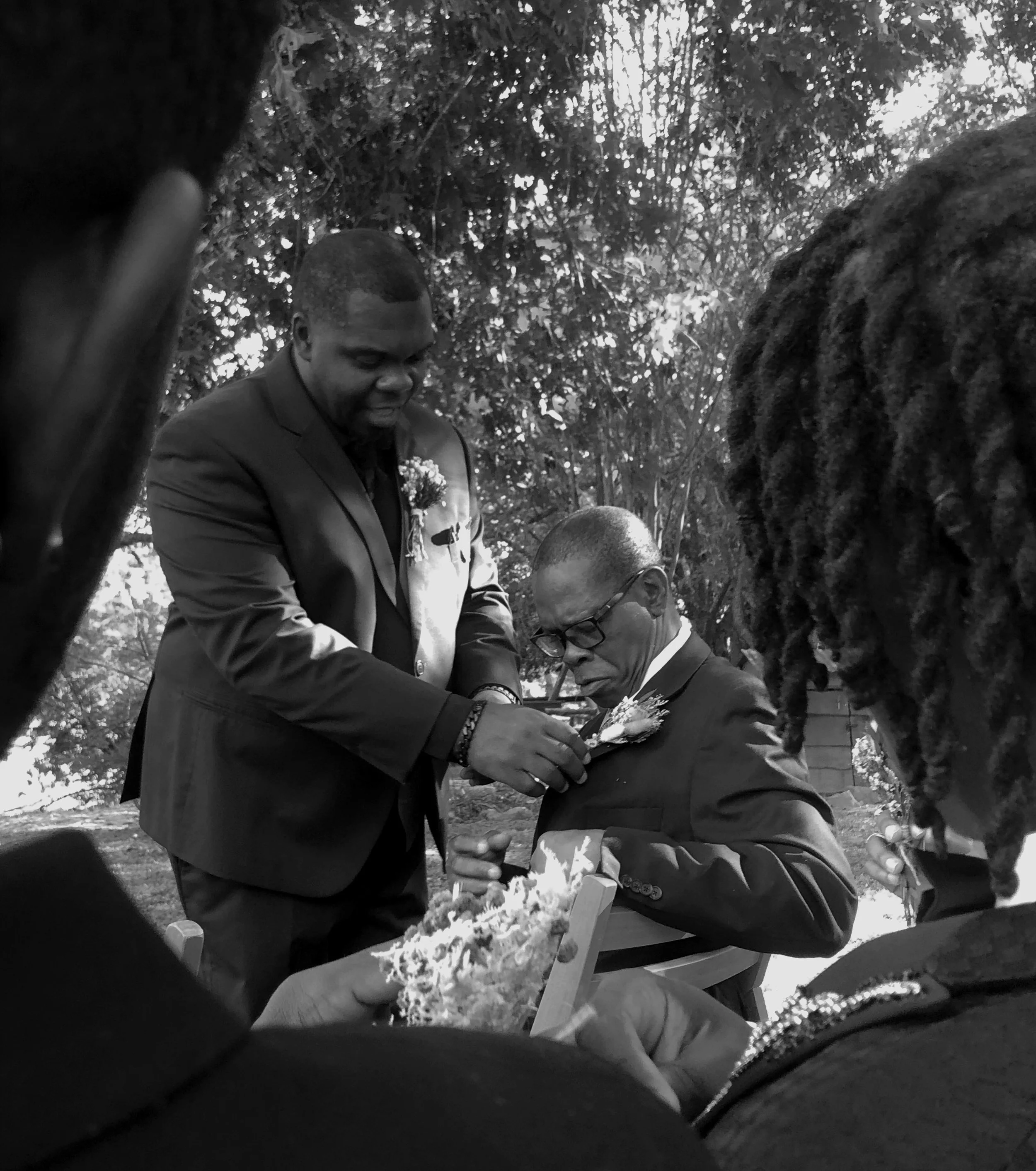 A black-and-white photo showing a man in a suit pinning a boutonniere on another man’s lapel during a wedding ceremony, with women partially visible in the foreground, outdoors with trees in the background.