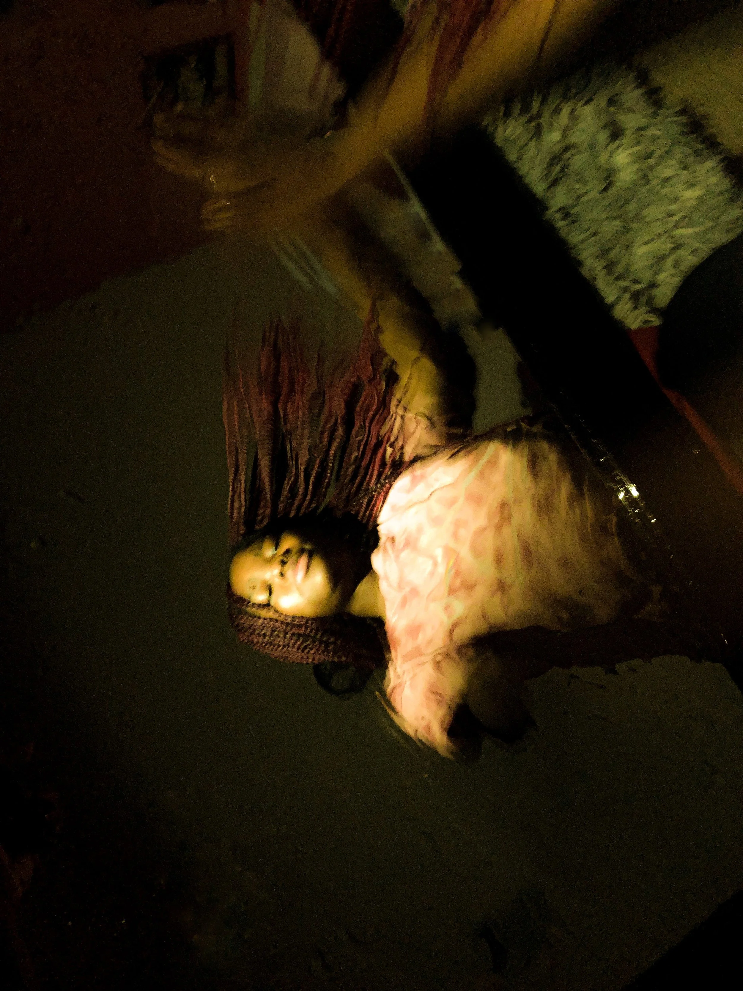 Woman lying on the floor in a dimly lit room with a striped carpet, wearing a pink top and patterned pants, with a small container nearby.