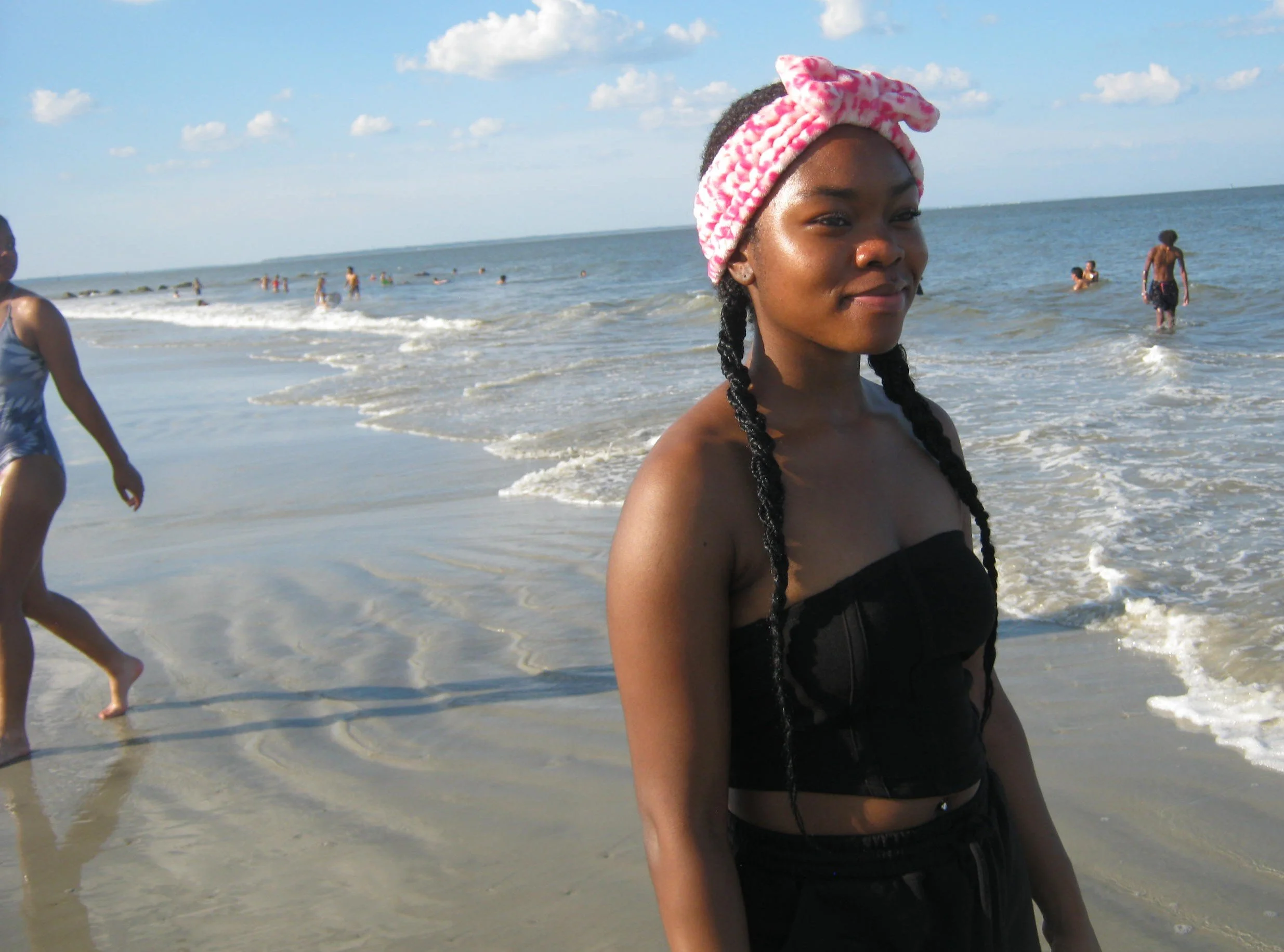 A young woman with braids and a pink headband standing at the beach with ocean waves and other people swimming in the background. Beach photography. Digital camera photography