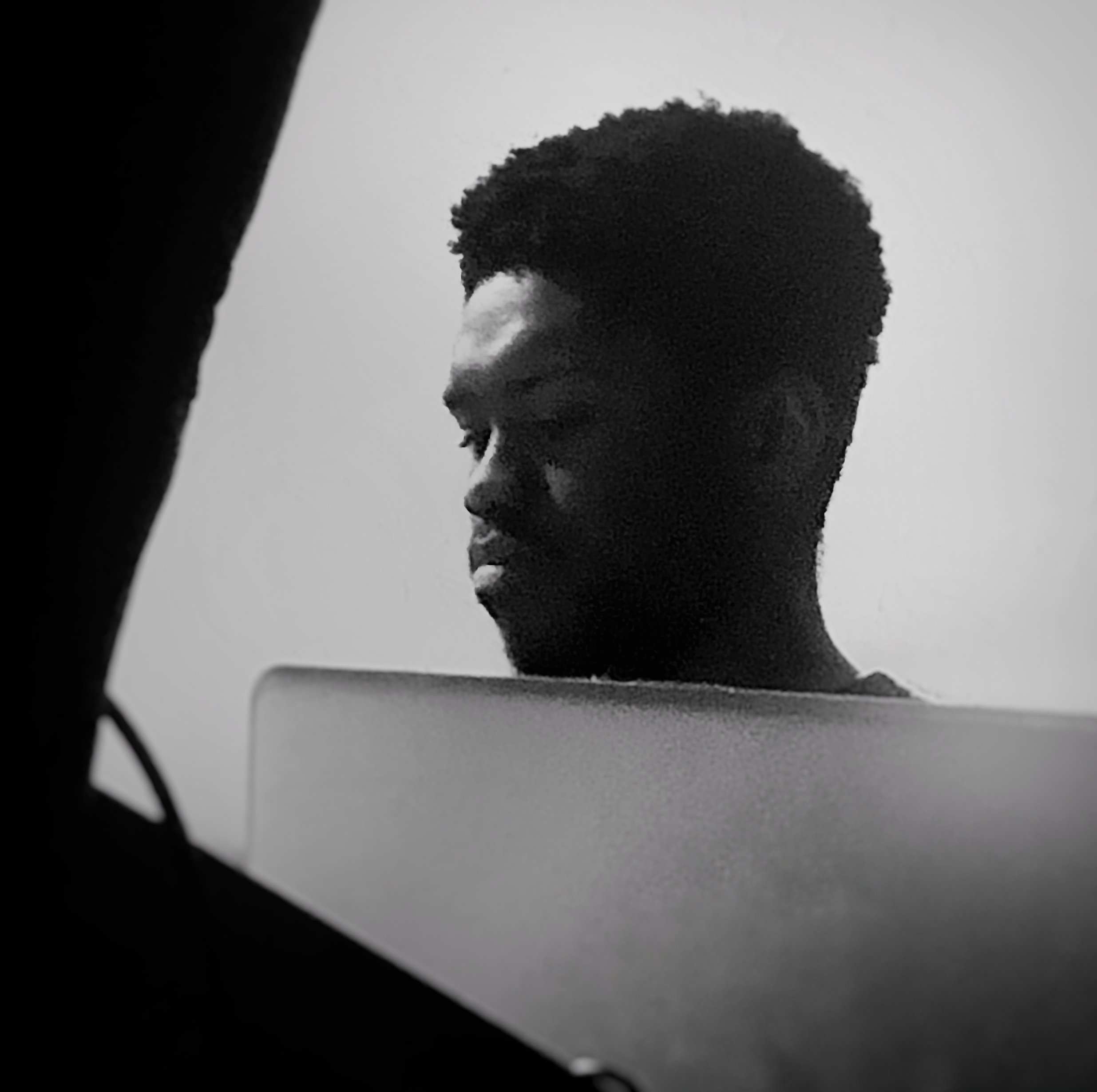 A black and white photo of a person with short, curly hair, working on a laptop, with part of a monitor visible in the foreground.