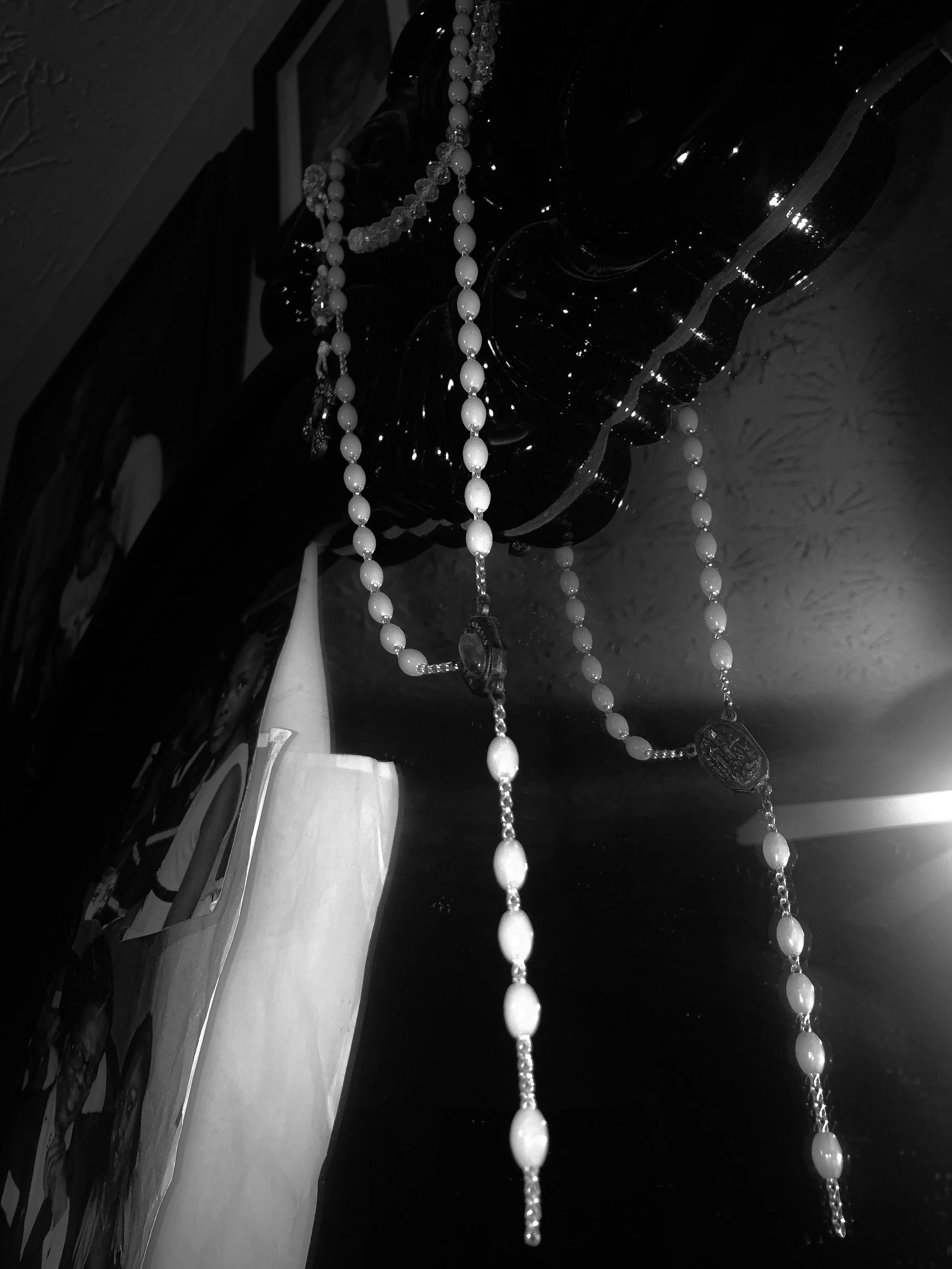 Black and white photo of hanging pearl necklaces, displayed on a dark decorative piece, with a light-colored box or book partially visible on the left side.