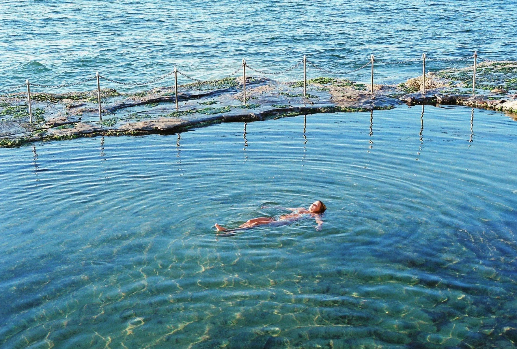 A woman floating on her back in a clear outdoor pool near the ocean, with a rocky barrier and chain fence in the background.